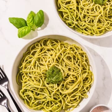 Two white bowls filled with pesto covered spaghetti and a dollop of pesto in the middle. Placed around the bowls there are two forks, basil for garnish, and salt.