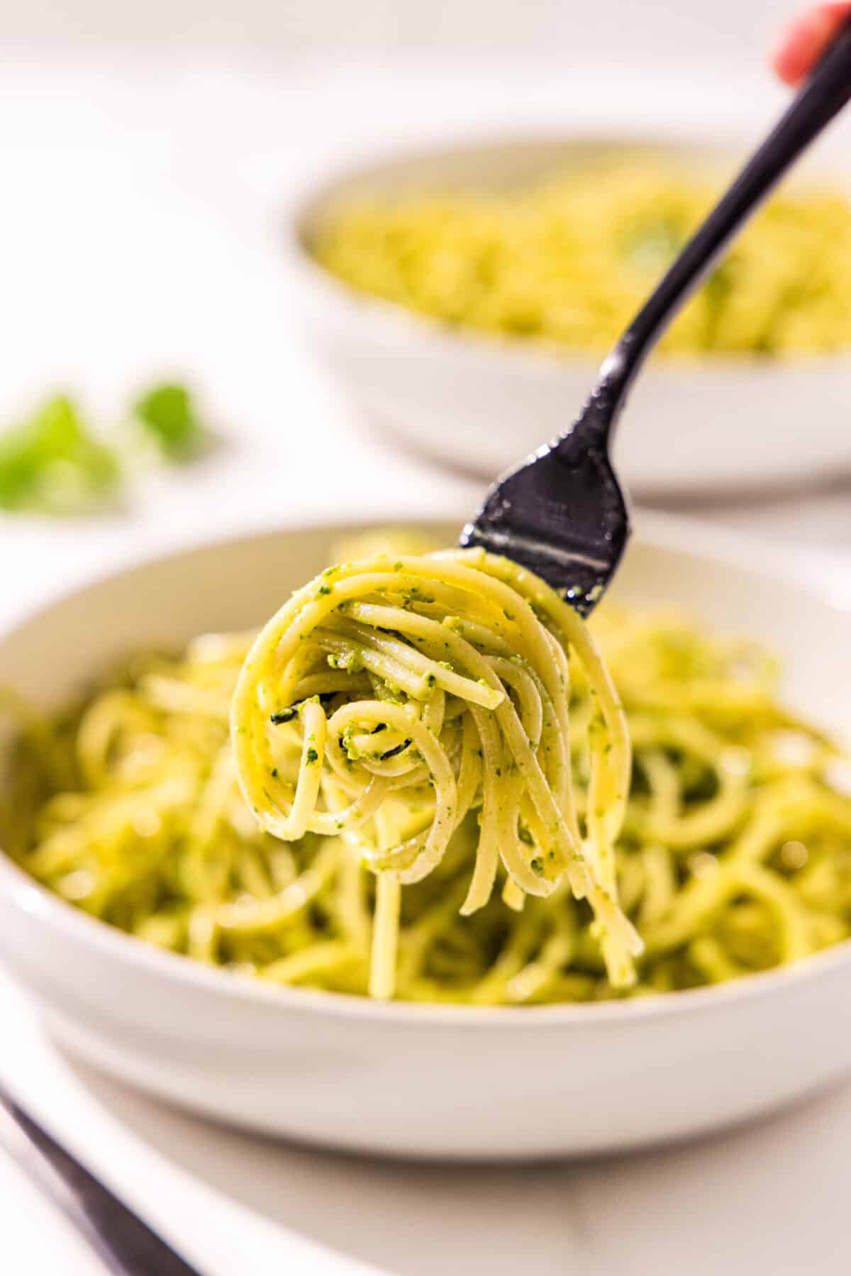 A white bowl filled with spaghetti pesto pasta with a bite of spaghetti on a black fork in the center of the bowl.