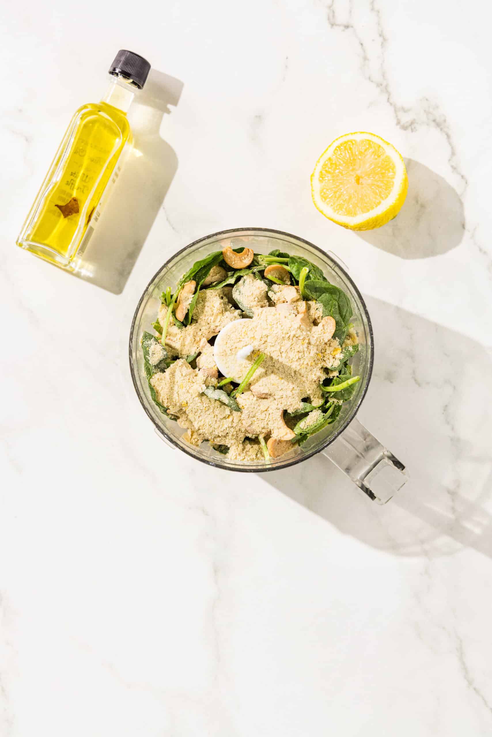 Food processor filled with spinach, basil, cashews, garlic, nutritional yeast, lemon juice, olive oil, and salt. Placed around the food processor is a small bottle of olive oil and half a lemon.