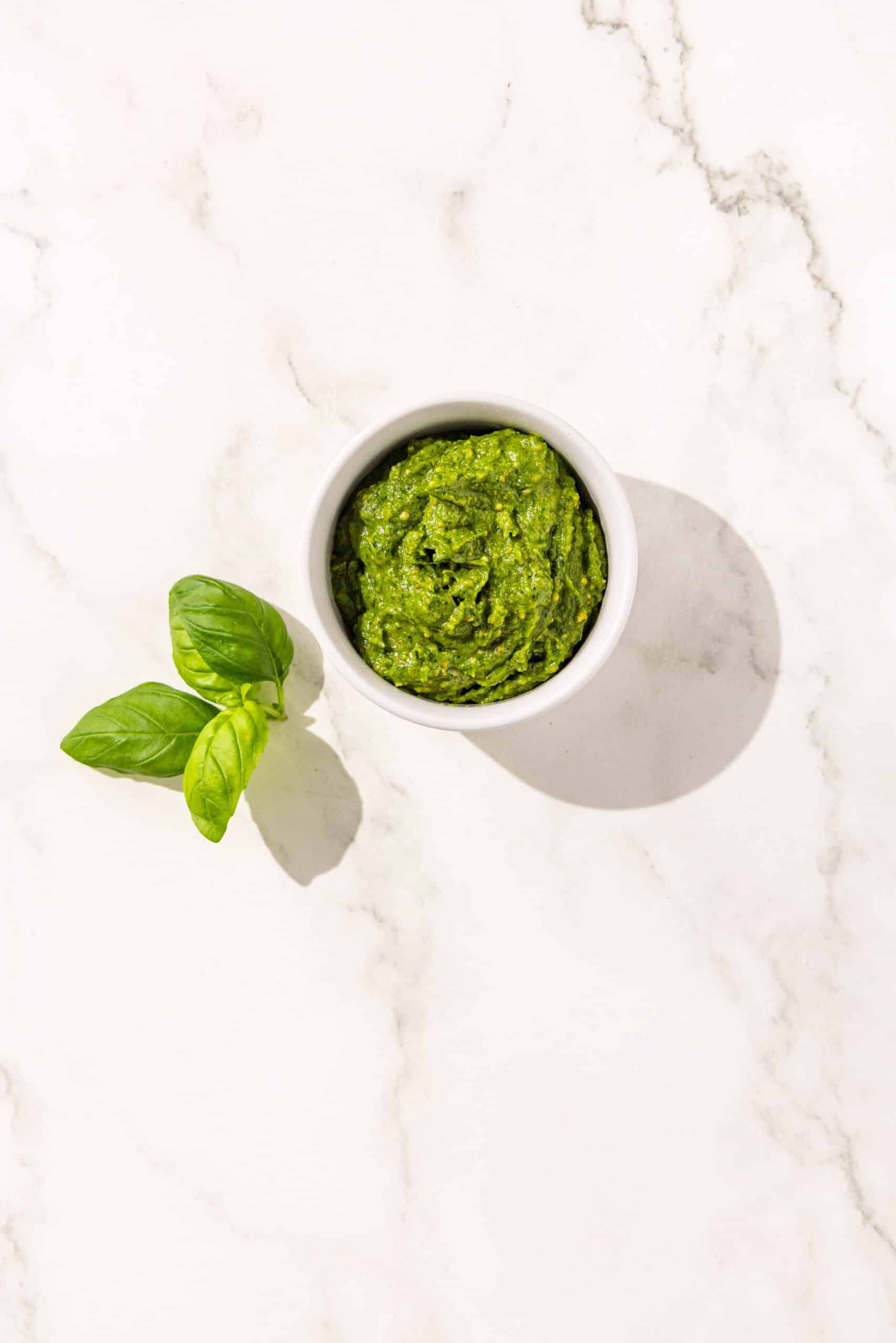 Small white bowl filled with pesto sauce. Beside the bowl is basil for garnish.