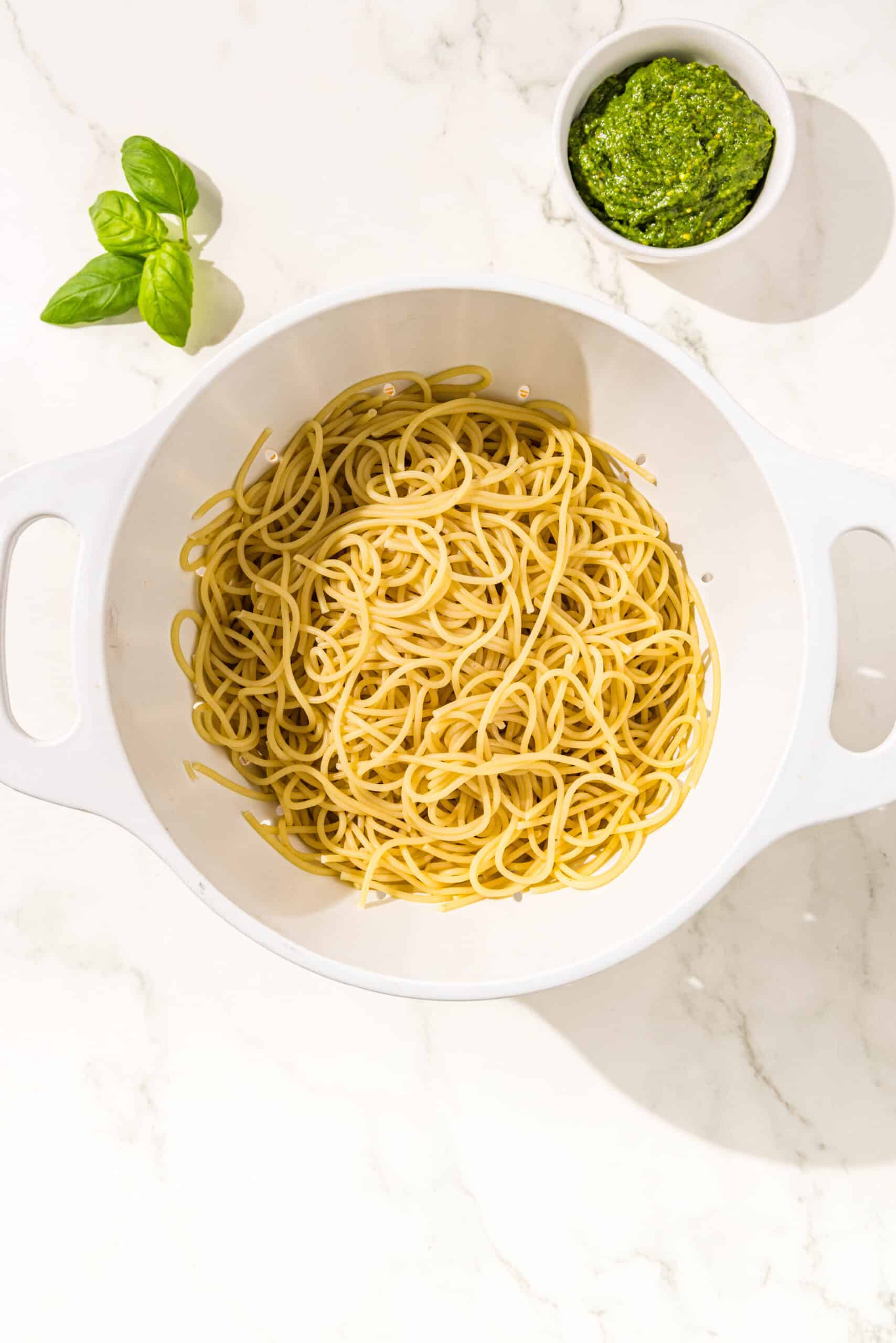 White colander filled with boiled spaghetti noodles. Around the colander is a small bowl of pesto sauce and basil.