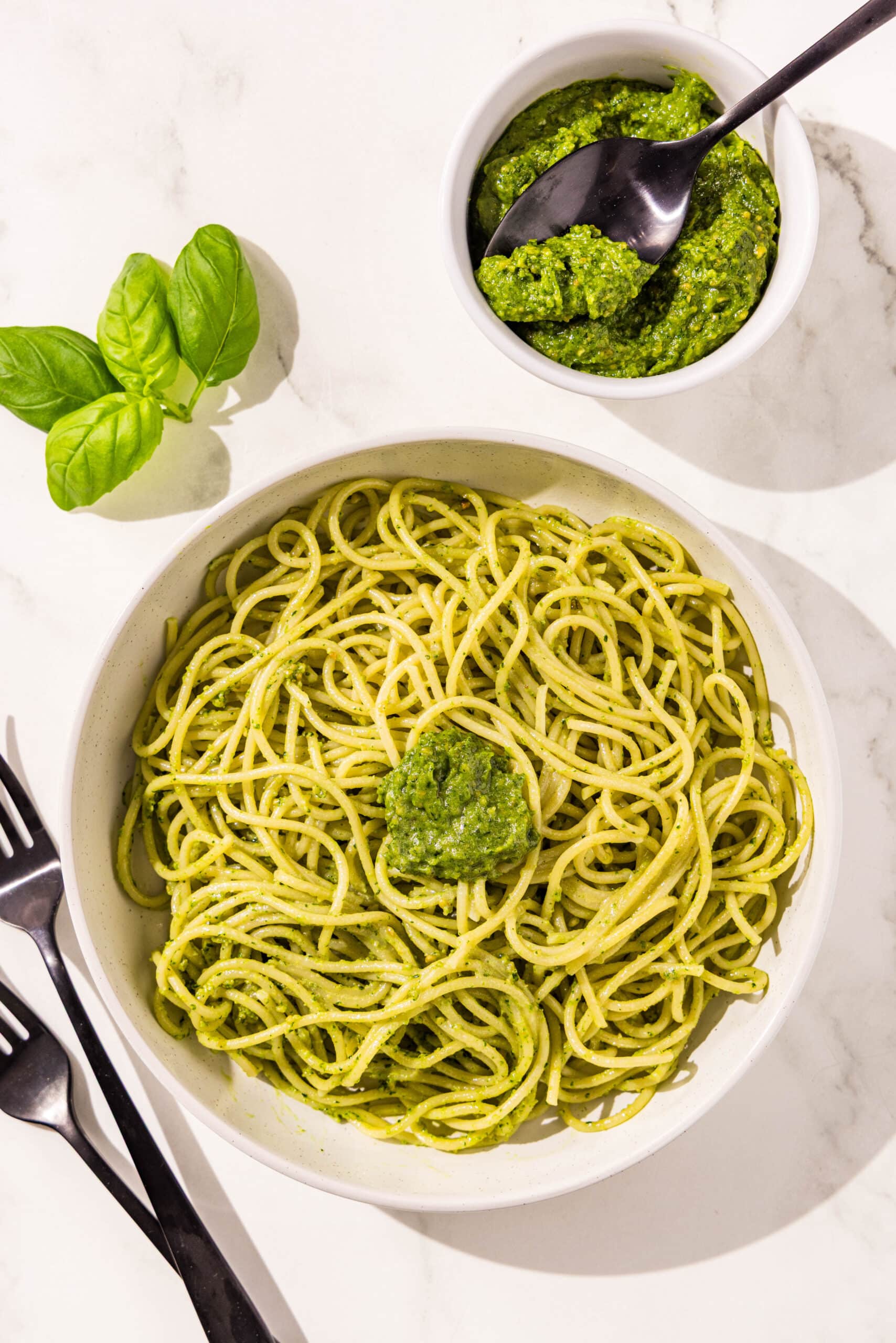 Big white bowl filled with pesto spaghetti and a dollop of pesto in the center. Around the bowl is two black forks, a small bowl of pesto sauce with a black spoon, and basil.