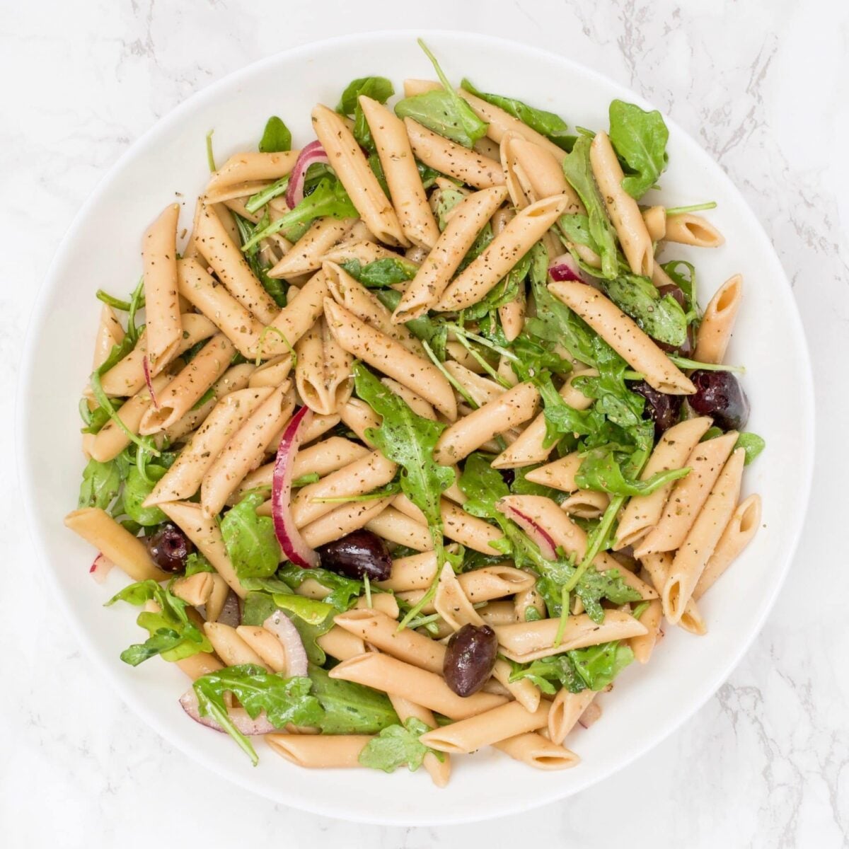 A bowl of pasta salad with arugula, sliced red onion, and kalamata olives in it.