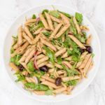 A bowl of pasta salad with arugula, sliced red onion, and kalamata olives in it.
