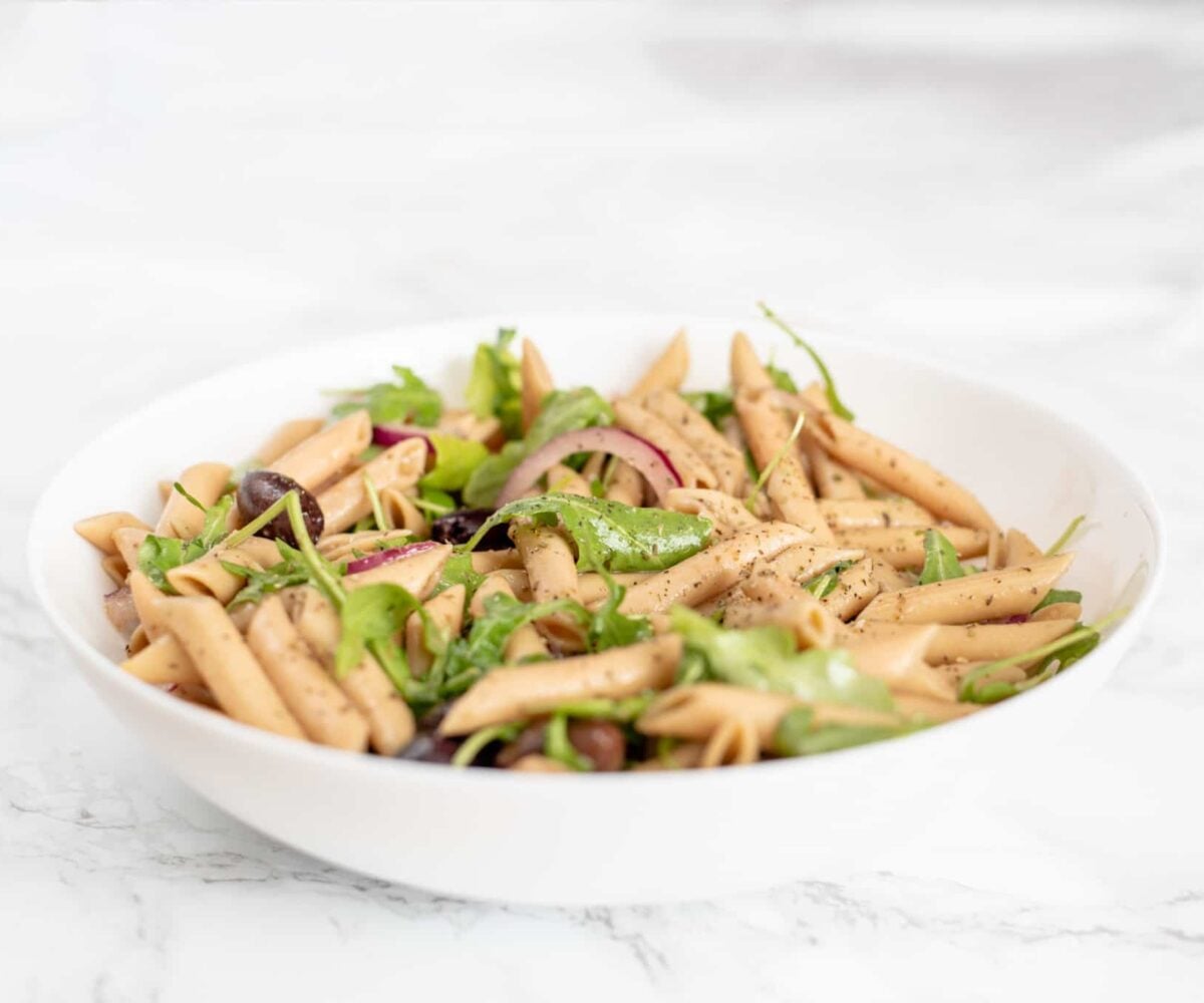 A bowl of pasta salad with arugula, sliced red onion, and kalamata olives in it.