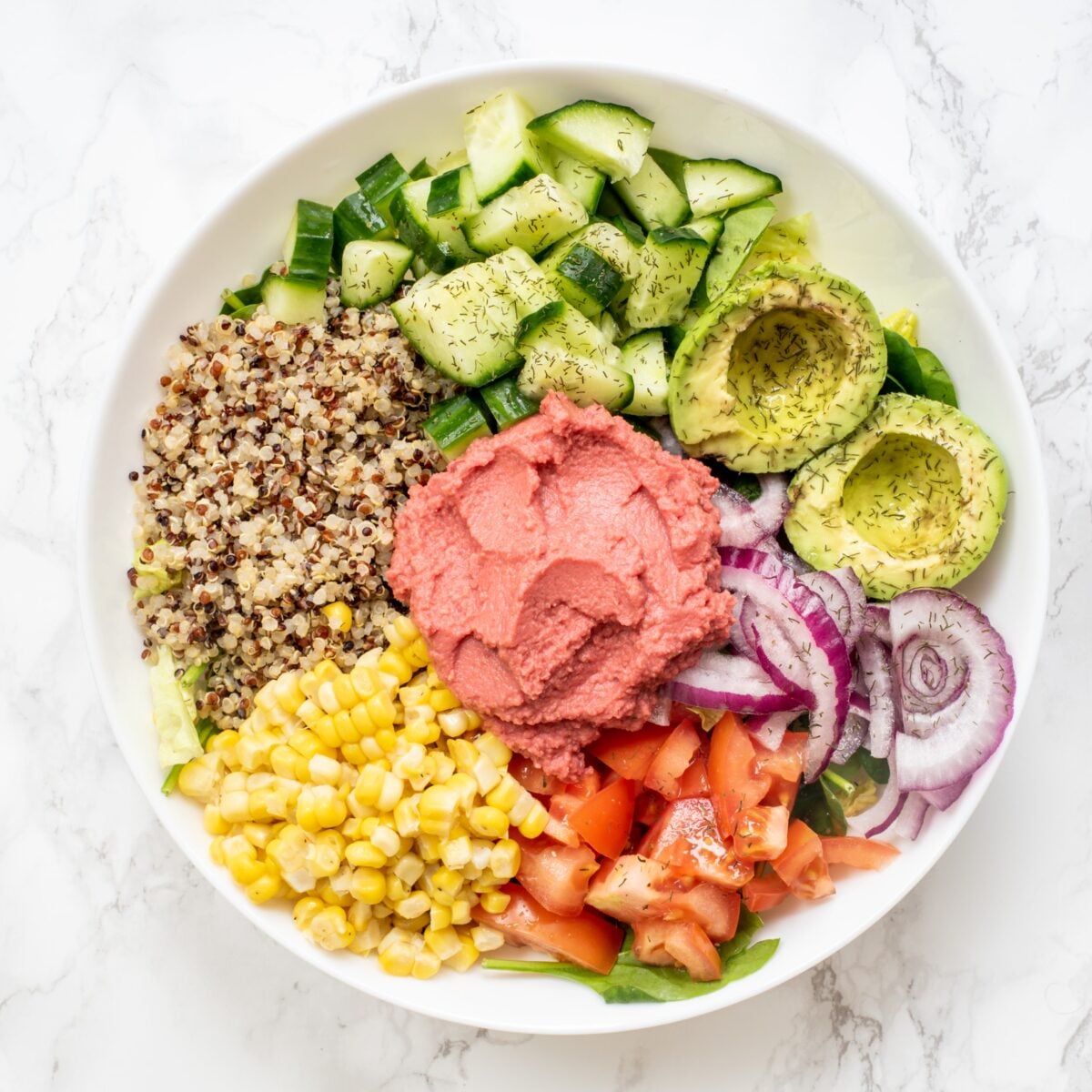 A white bowl filled with piles of spinach, quinoa, diced cucumber, halved avocado, red onion sliced, tomatoes, and corn with a scoop of pink hummus in the center.
