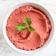 A white bowl of beet hummus topped with a cilantro leaf in the center.