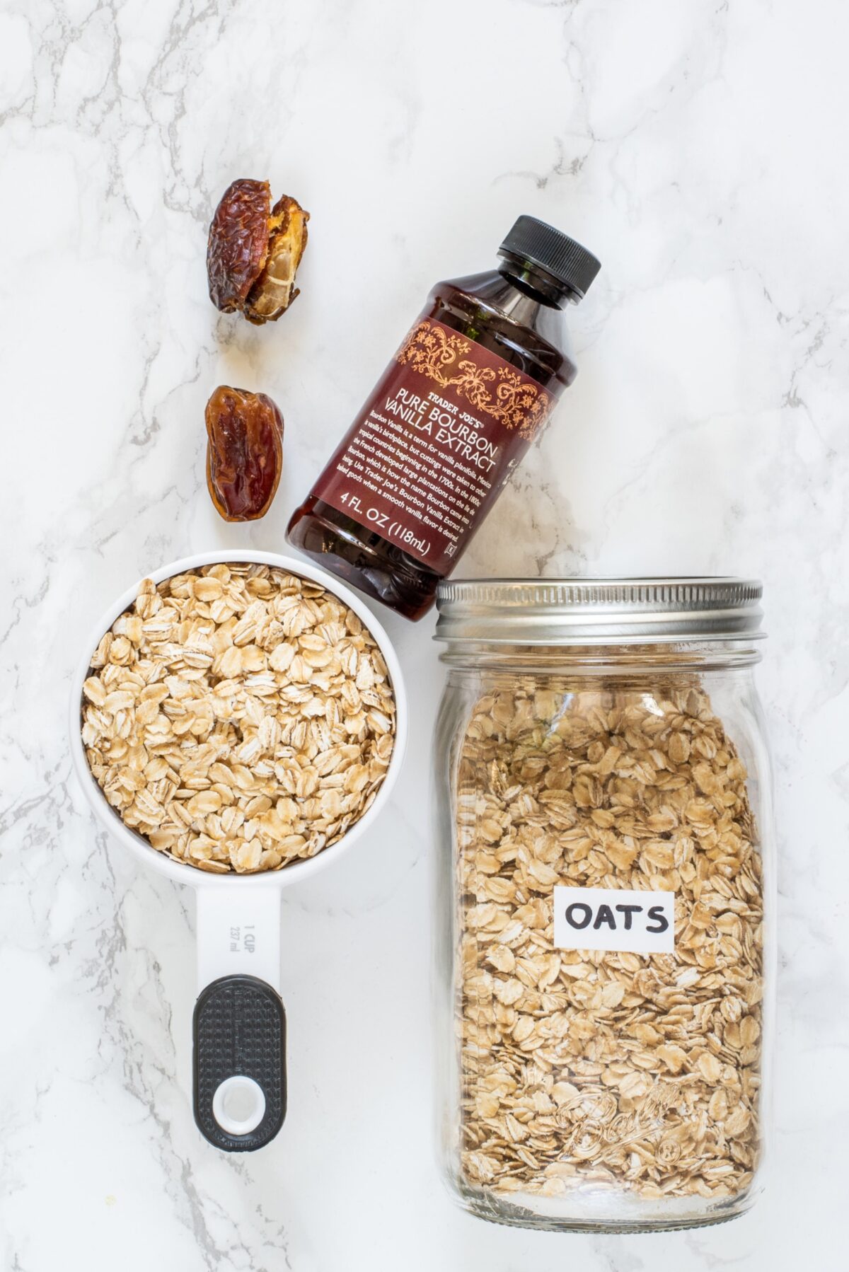 A jar of oats, a 1 cup measuring cup filled with oats, a bottle of vanilla extract, and 2 medjool dates.