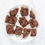A white round plate of 10 misshapen vegan chickpea brownies topped with chocolate chips.