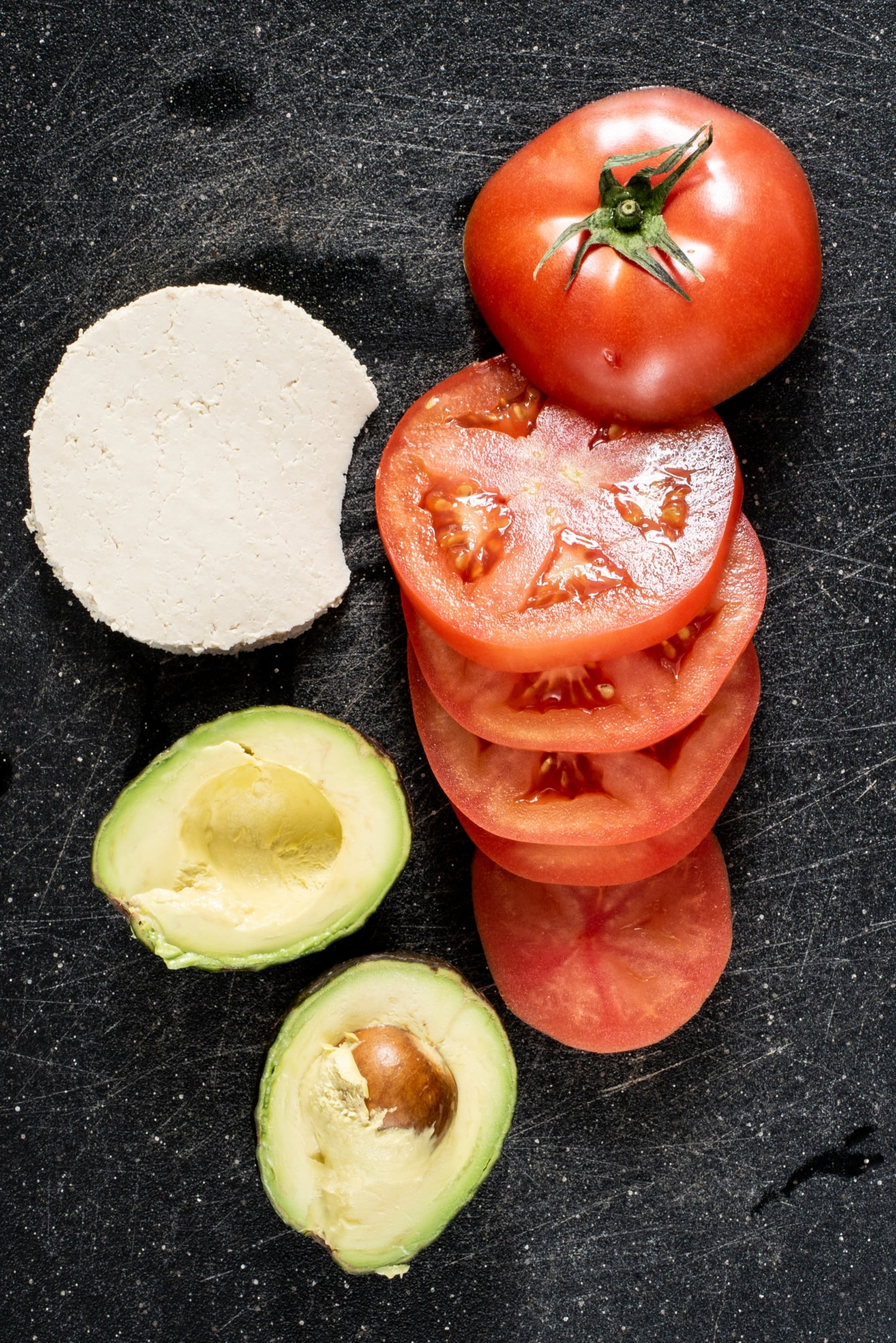 A cutting board with tofu, tomato slices, and an avocado.