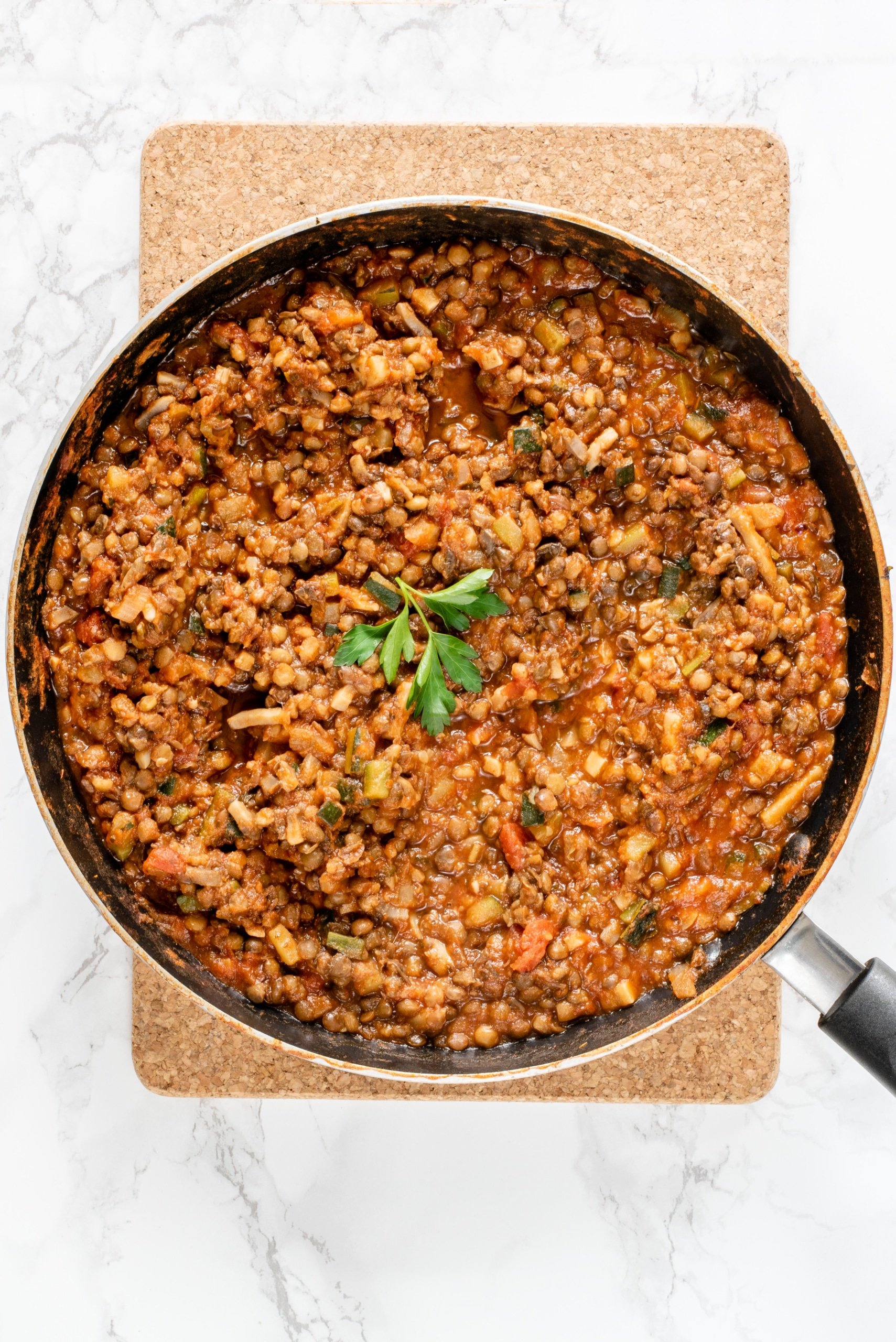 A large black frying pan on a cork board with cooked lentil bolognese in it and a sprig of parsley in the center.