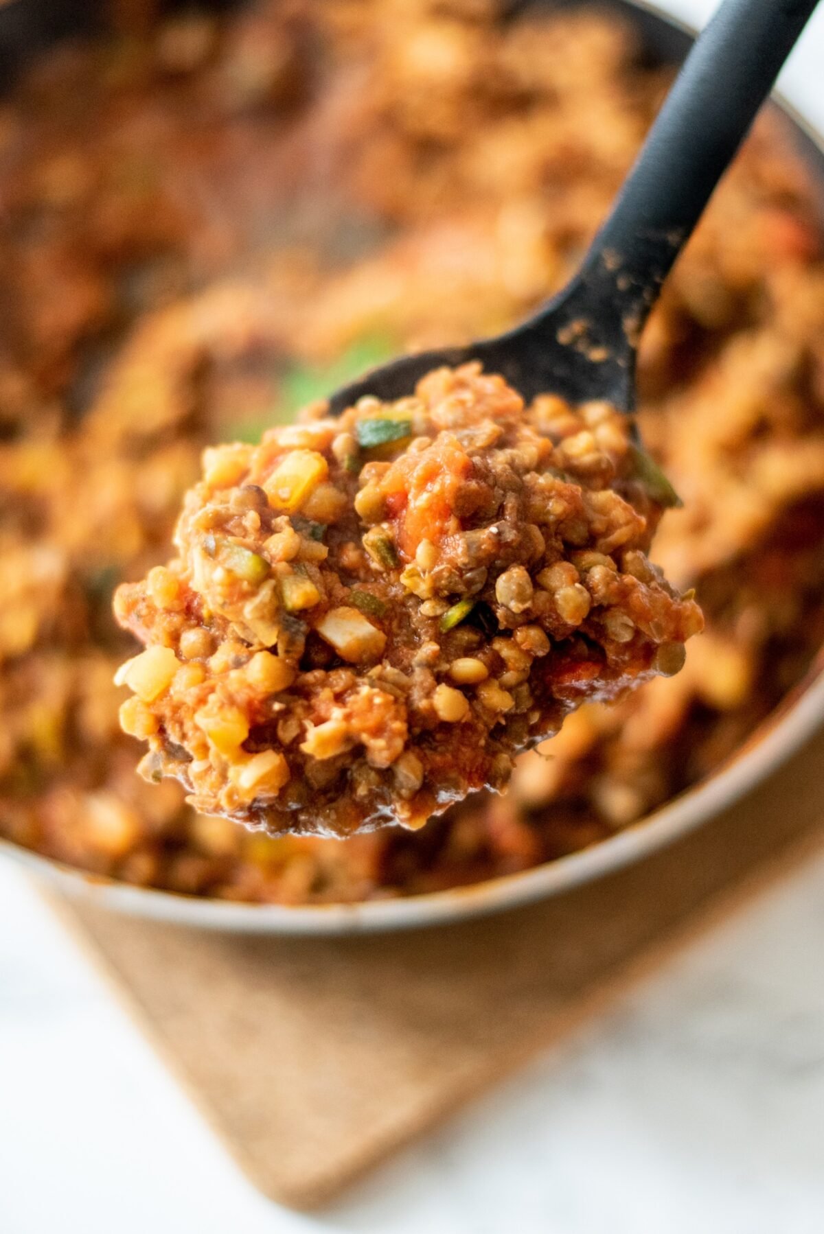 A black spoon holding a scoop of vegan lentil bolognese sauce over a pan with remaining sauce.
