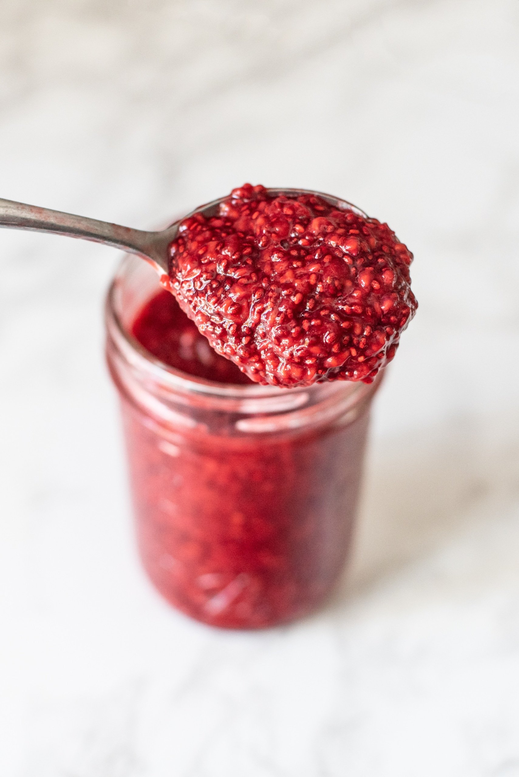 A glass mason jar filled with Raspberry Chia Jam with a spoon sticking out holding a scoop of the jam.