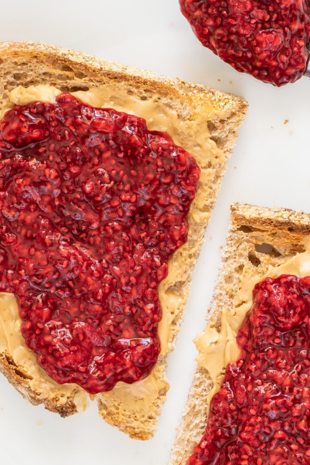 Close up of one slice of toast cut in half with almond butter and Raspberry Chia Jam on it and a scoop of raspberry chia jam on the side on the plate.