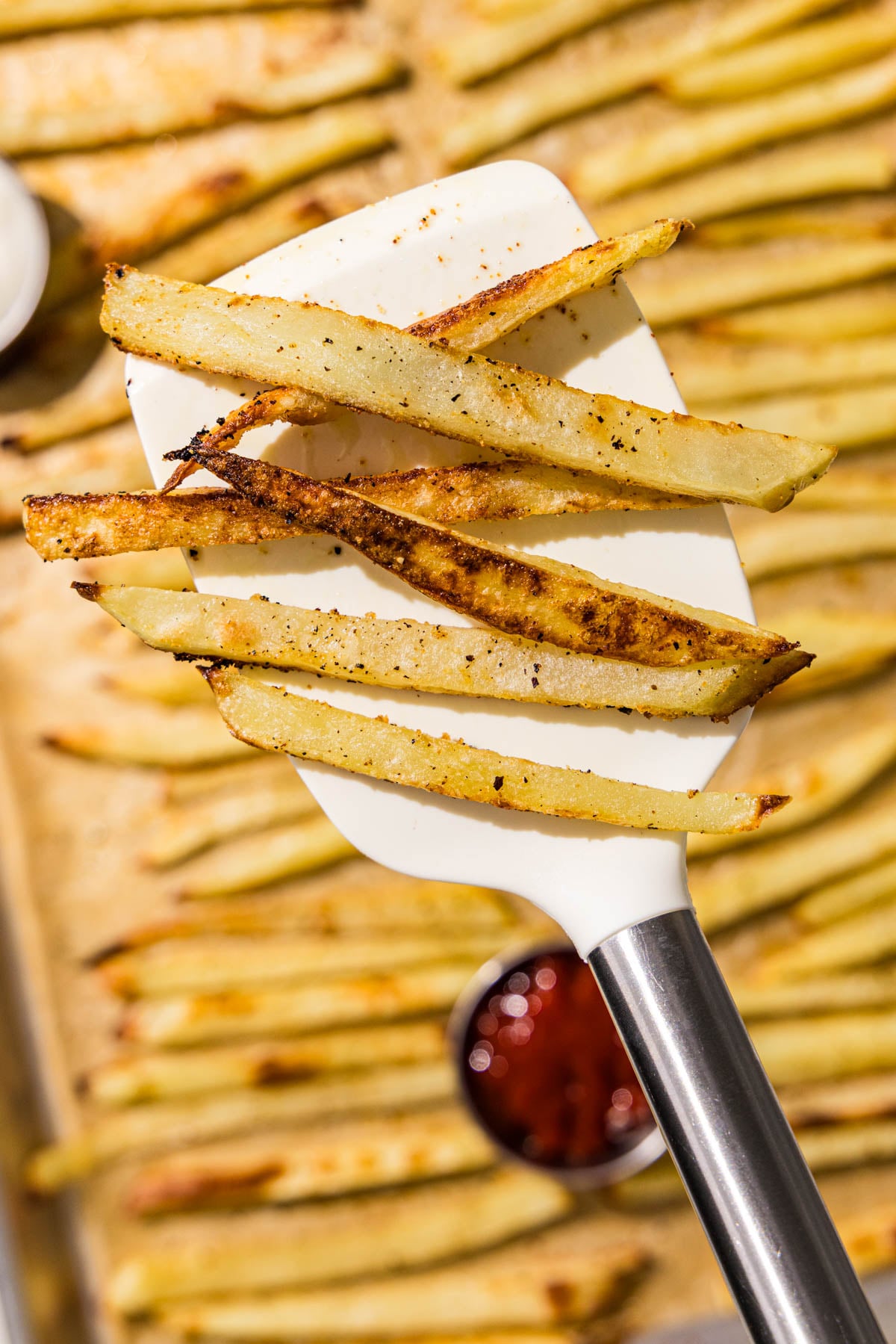 A white spatula holding 6 baked french fries over a sheet pan with remaining french fries and a small bowl of ketchup.