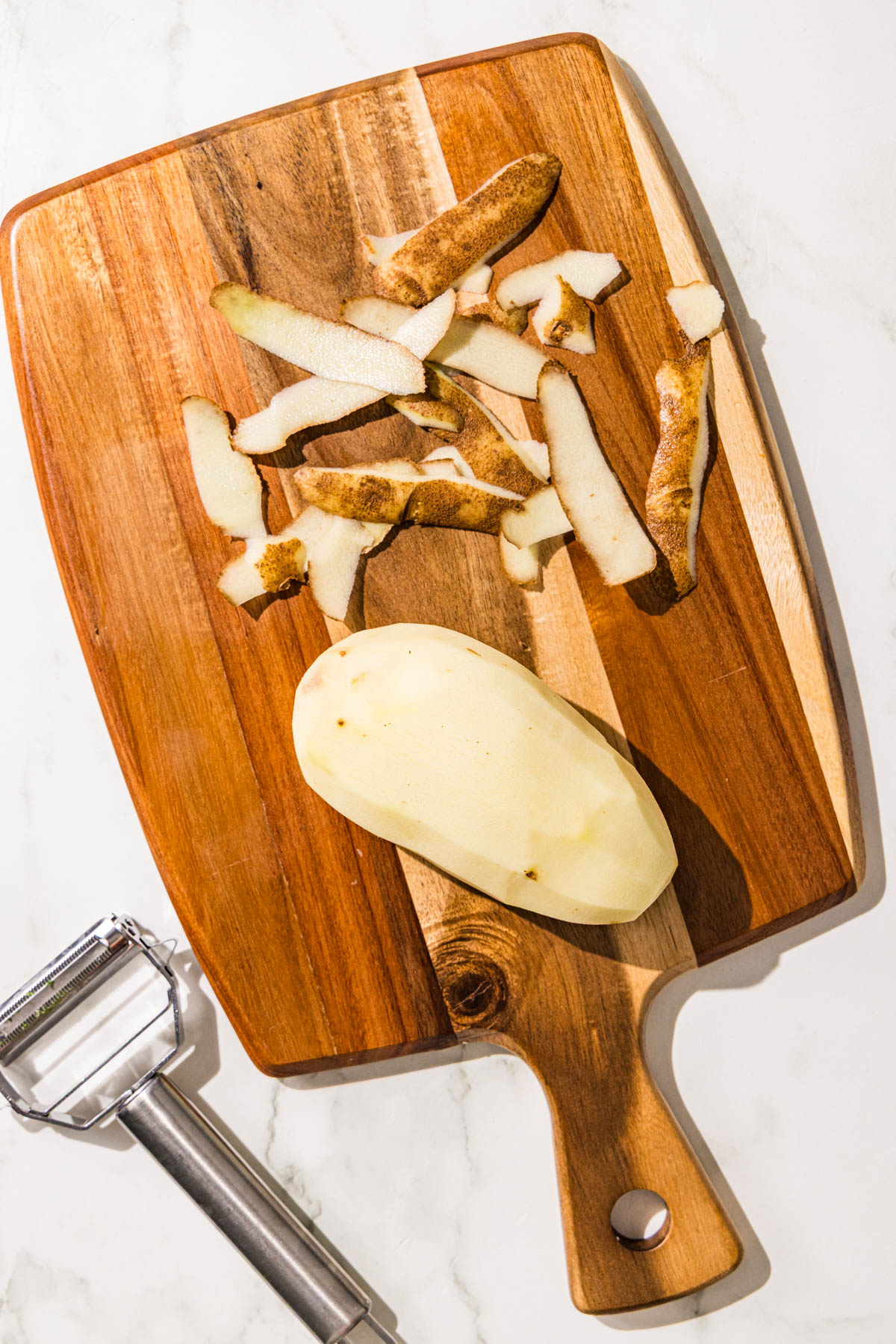 A brown wooden cutting board with a peeled potato and potato peels on it and a silver peeler.