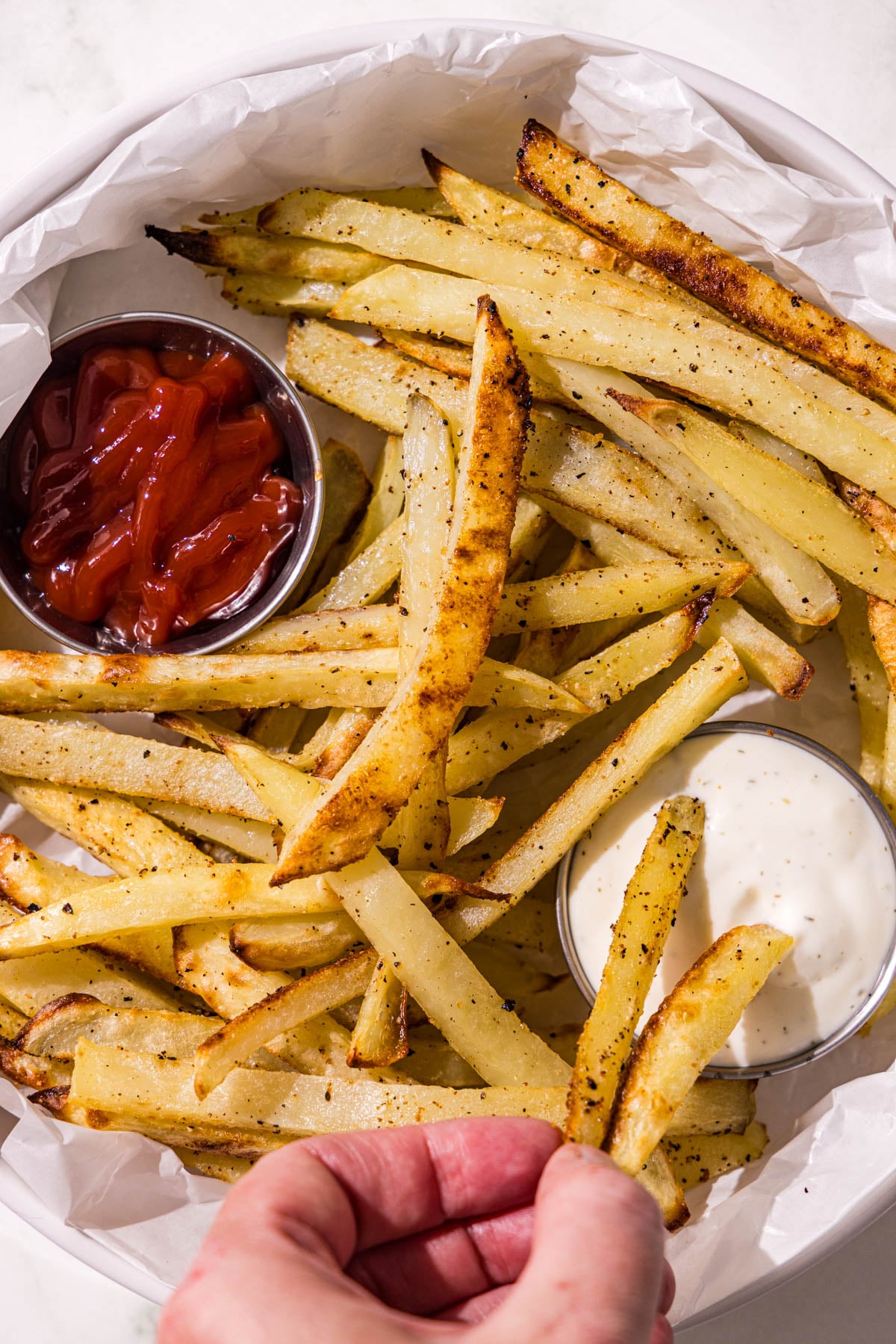 A white bowl lined with white parchment paper filled with baked french fries and two metal containers of ketchup and ranch.