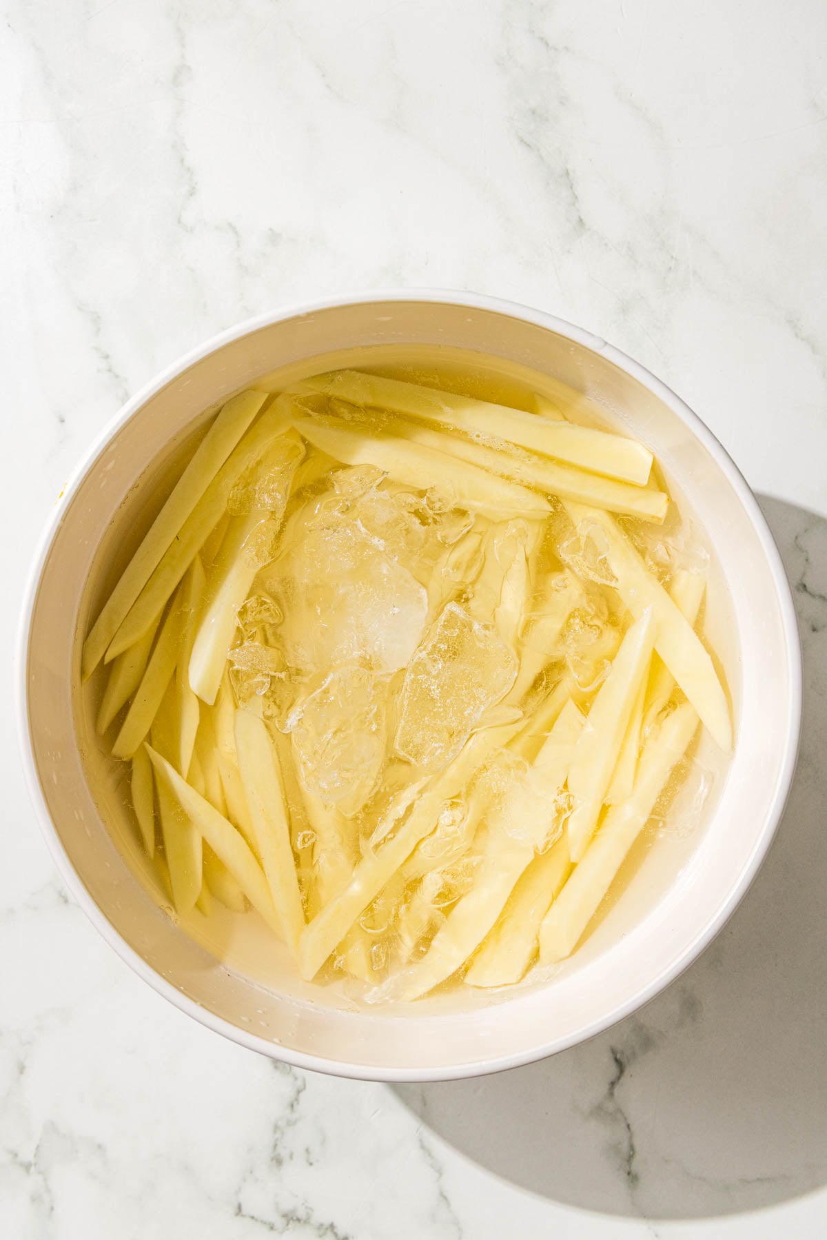 A large white glass bowl filled with ice water and sliced potatoes.