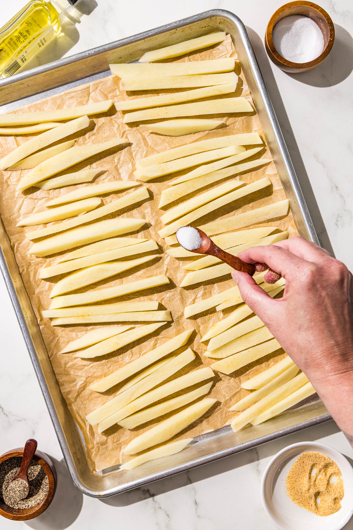 A large sheet pan lined with parchment paper and topped with sliced potato fries. Beside the pan is a dish of salt, pepper, garlic powder, and a small bottle of oil.