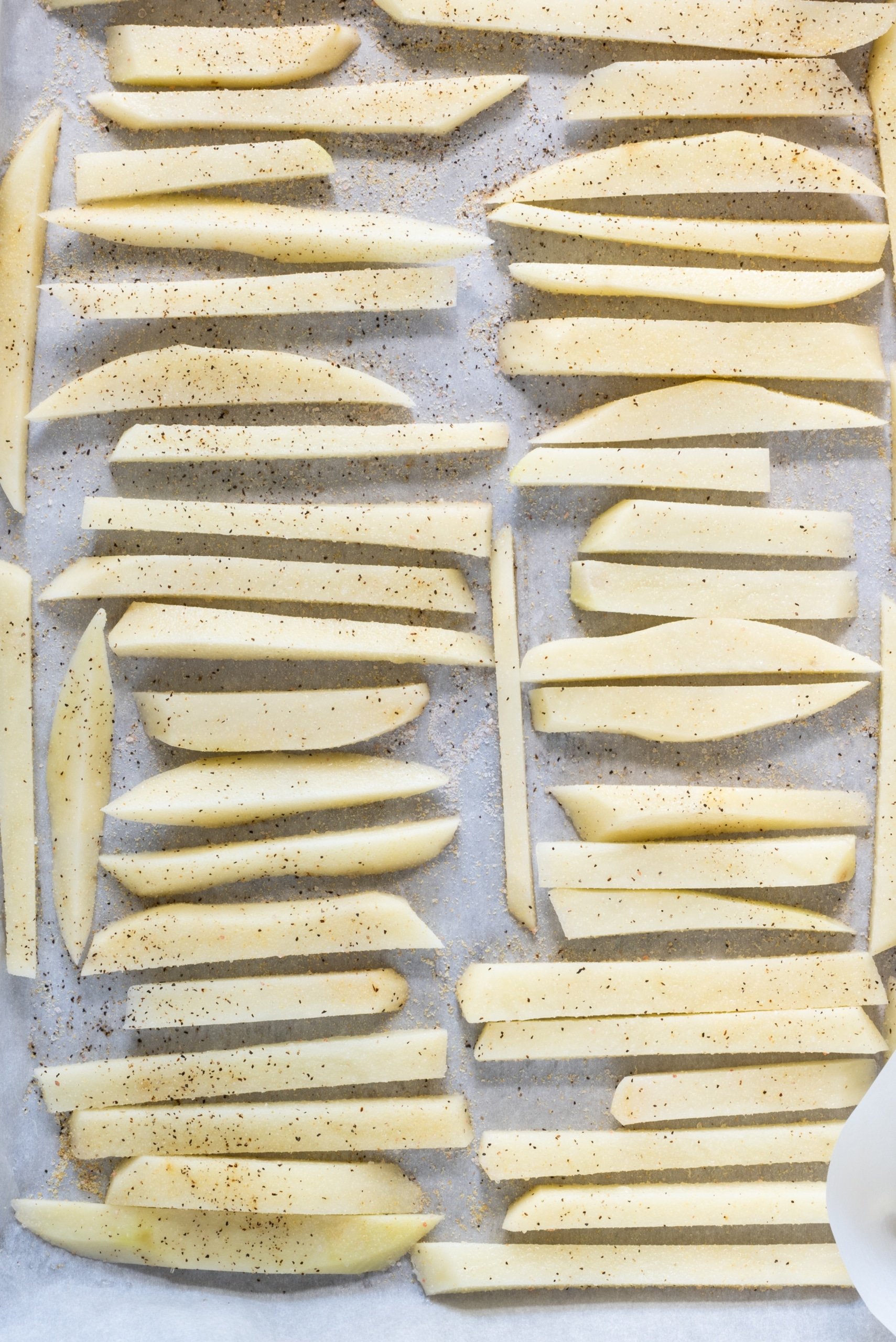 A pan lined with parchment paper with potato fries lined in single file, ready to be baked.