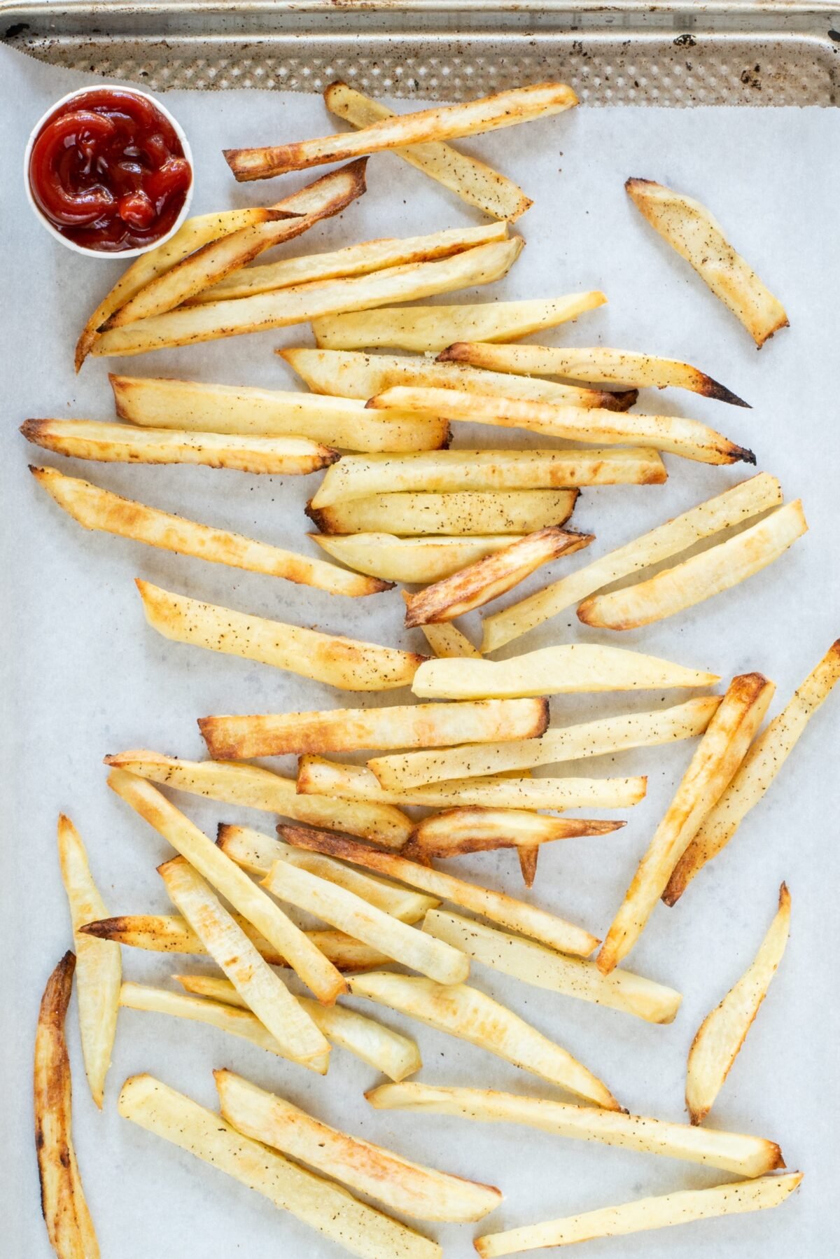 Crispy Baked French Fries on a baking sheet lined with parchment paper. In the top left corner of the pan is a small white cup of ketchup.