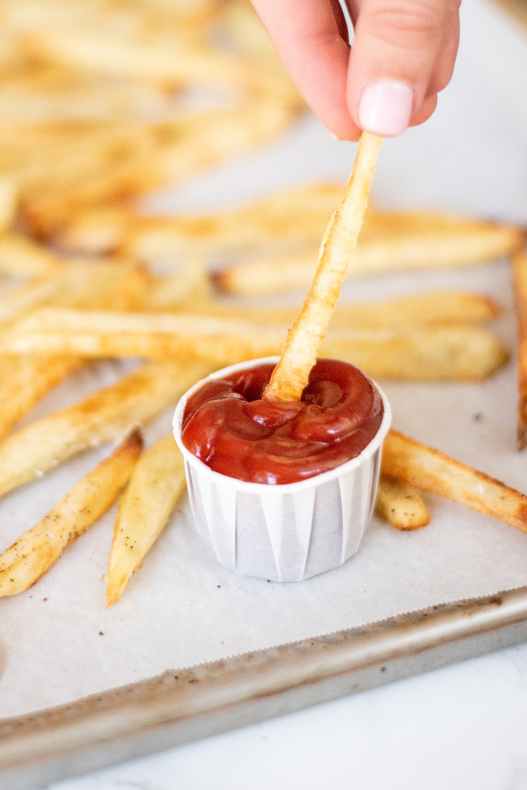 Crispy baked french fries on a pan lined with parchment paper. In the center is a small white cup of ketchup with a white hand dipping a fry into it.