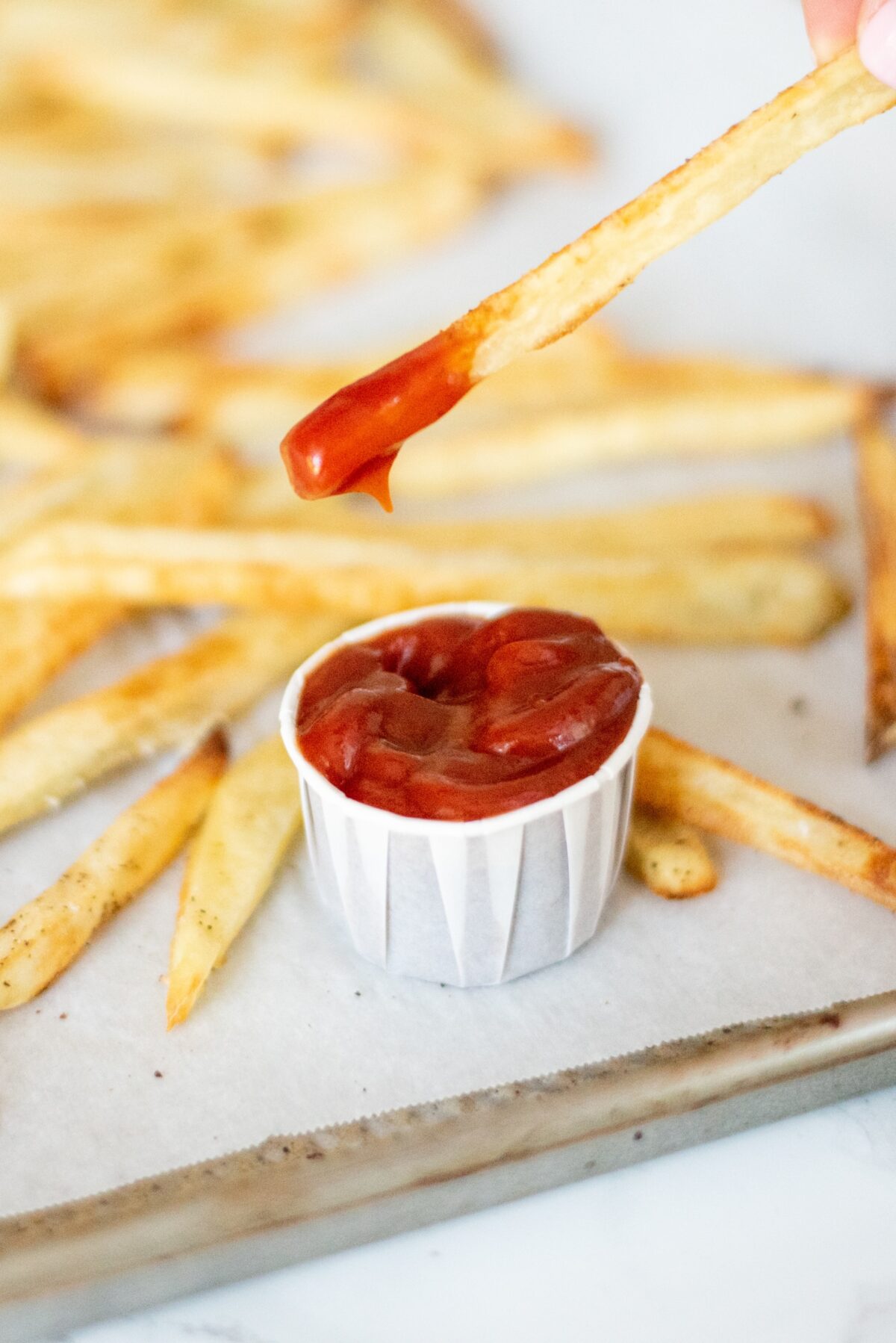 Crispy Baked French Fries on a pan lined with parchment paper. In the center of the pan is a small white cup of ketchup with a single fry being dipped into it.