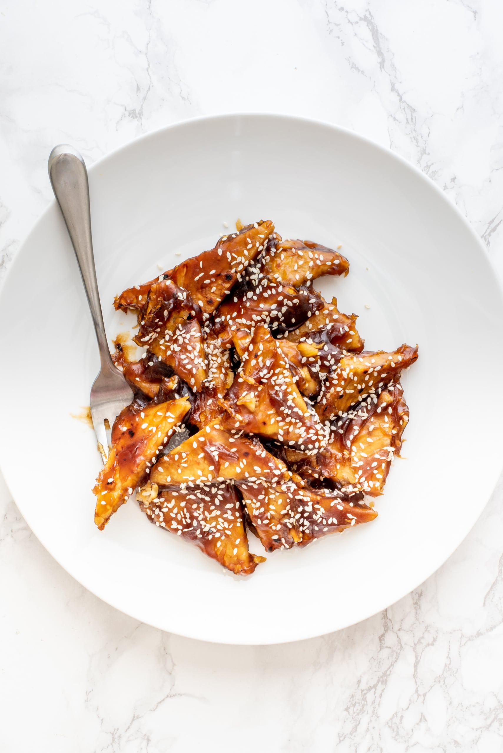 A white bowl filled with general tso's tempeh garnished with sesame seeds and a silver fork sticking out from the bowl.
