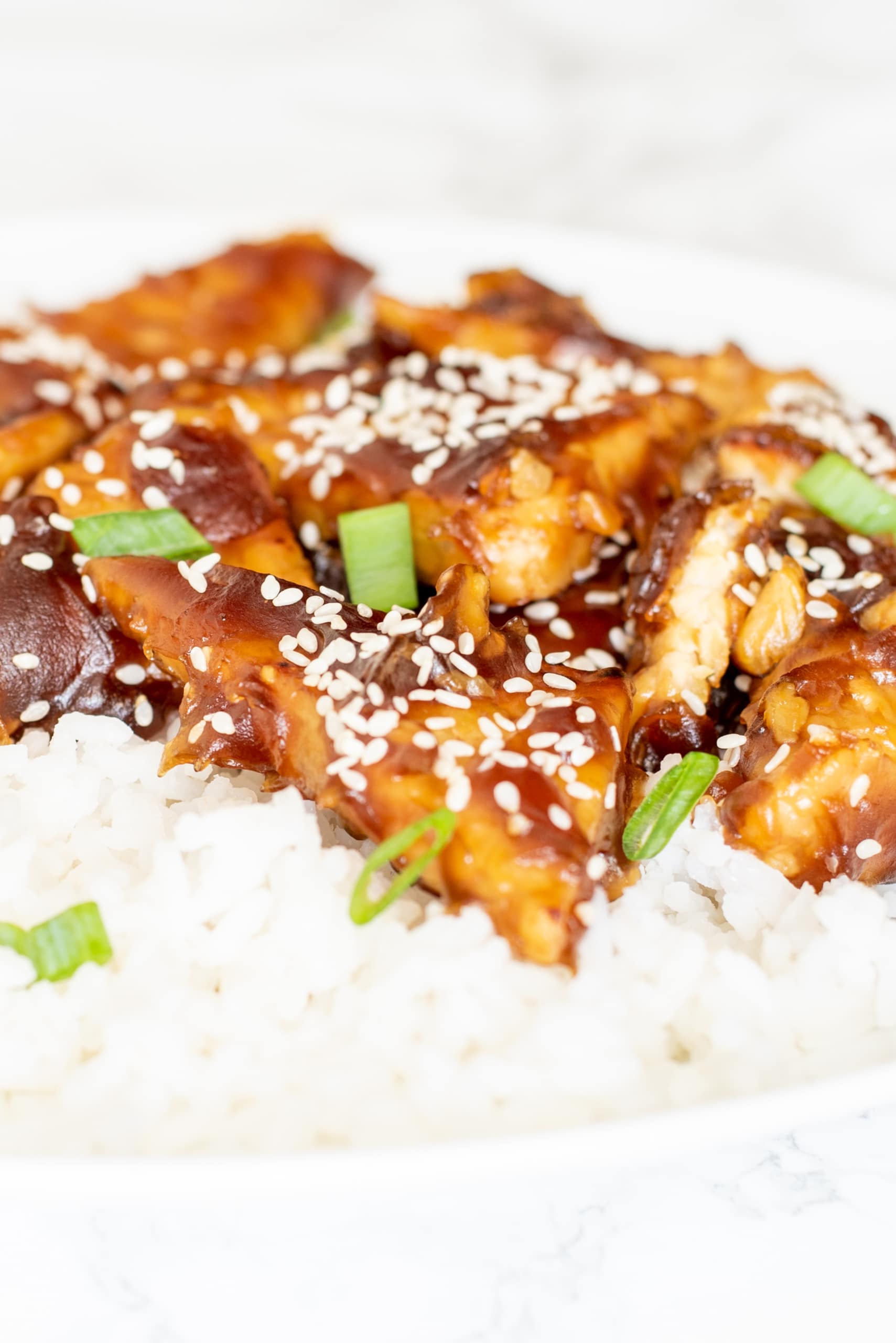 General Tso's Tempeh on a white plate on a bed of white rice garnished with sesame seeds and green onion.