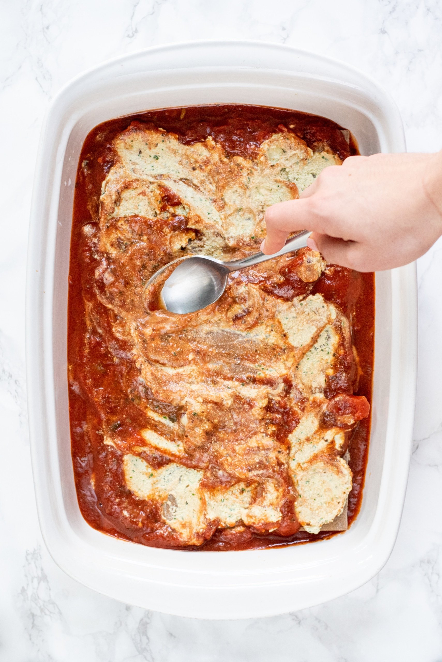 A 9x13 baking dish filled with marinara, lasagna noodles, and ricotta. A white hand is holding a silver spoon that is swirling around the sauce and ricotta.