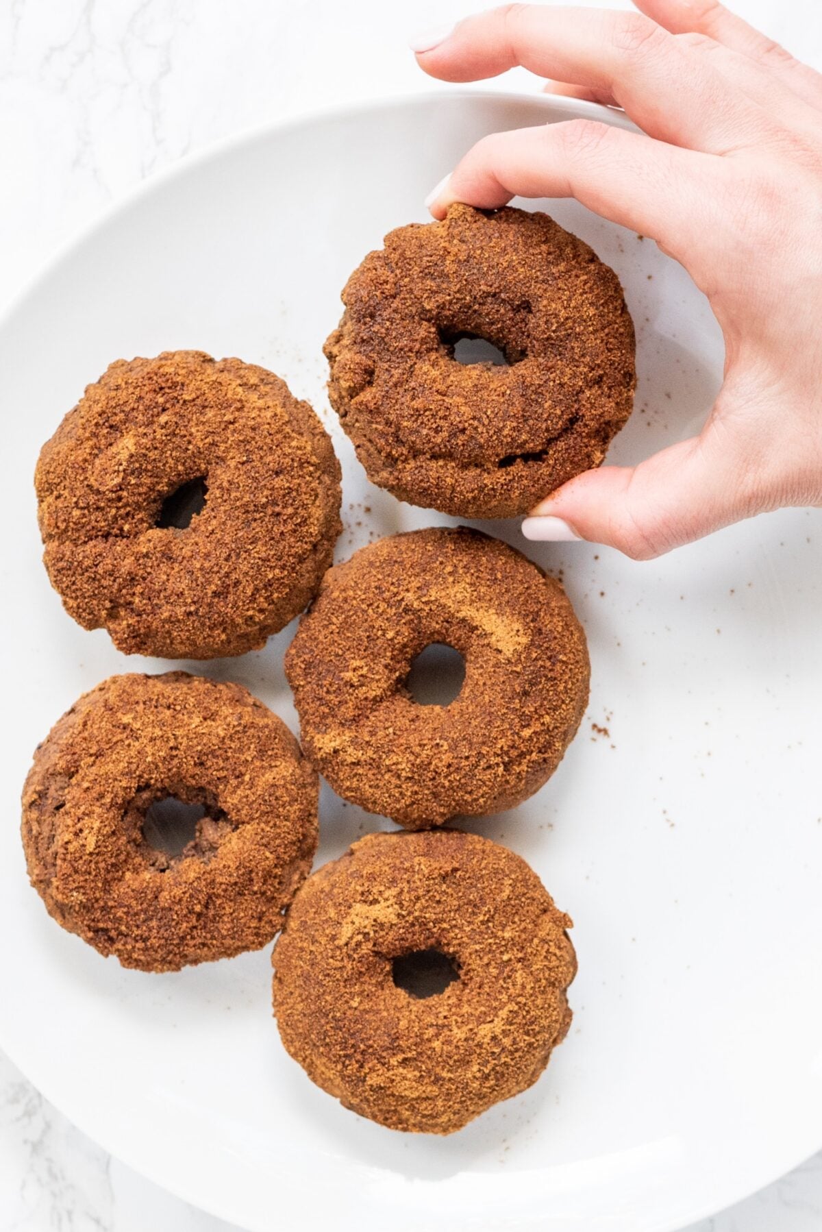 A white plate with 5 cinnamon sugar donuts on it and a white hand picking one up off the plate.