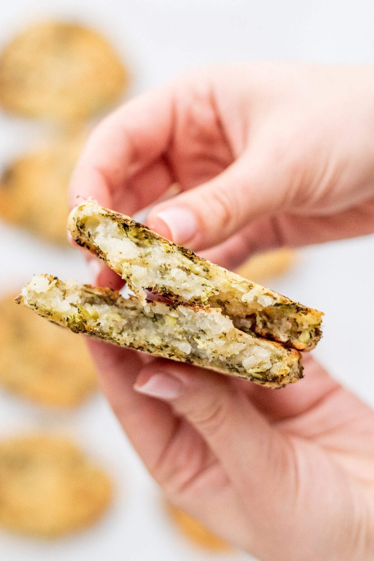 Two white hands holding a broccoli potato hashbrown broken in half.