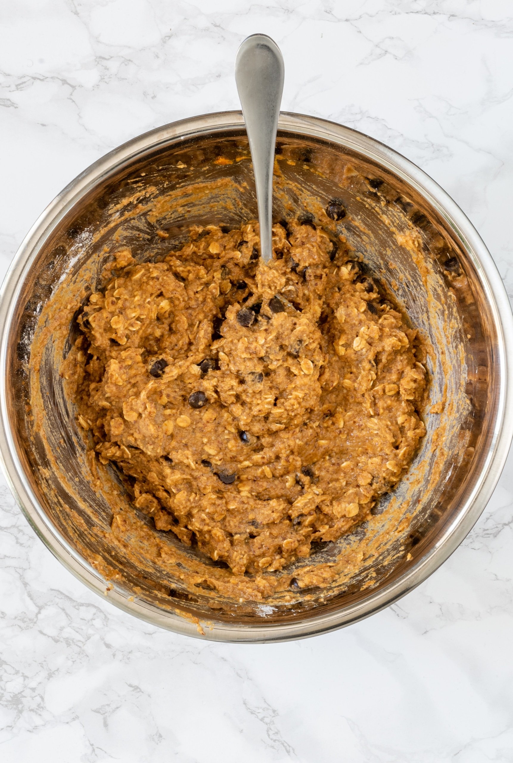 A silver bowl filled with an orange batter with chocolate chips and oats in it. In the bowl is a silver spoon.