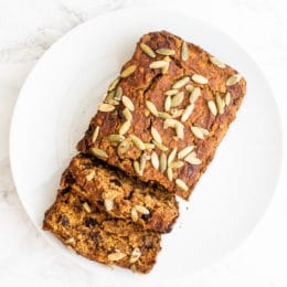 A loaf of pumpkin bread on a white plate with two slices cut off on the end. The loaf is topped with pumpkin seeds.
