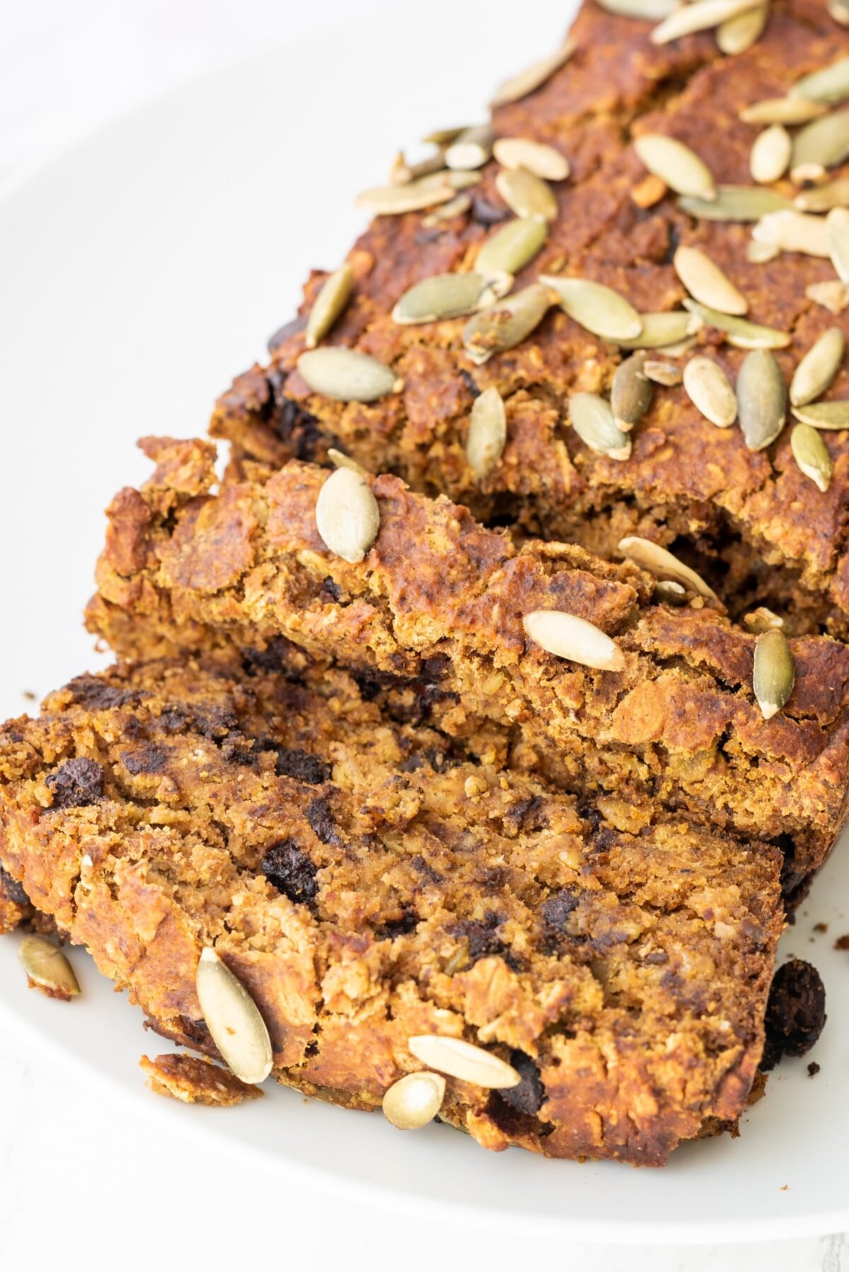 A close up of Vegan Chocolate Chip Pumpkin Bread topped with pumpkin seeds on a white plate with two slices cut off on the end.