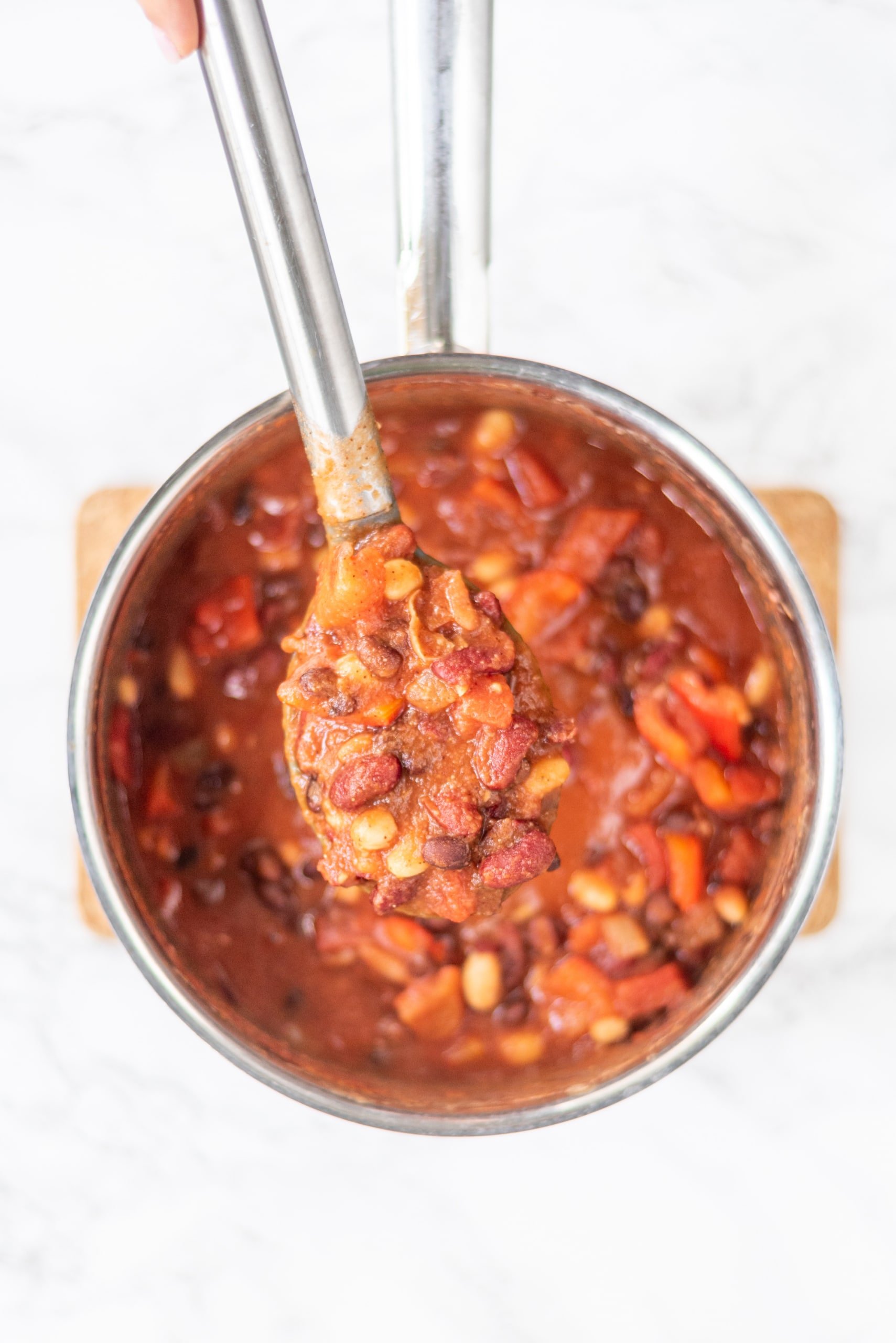A silver pot filled with chili. A silver spoon is held above the pot with the spoon filled with chili.