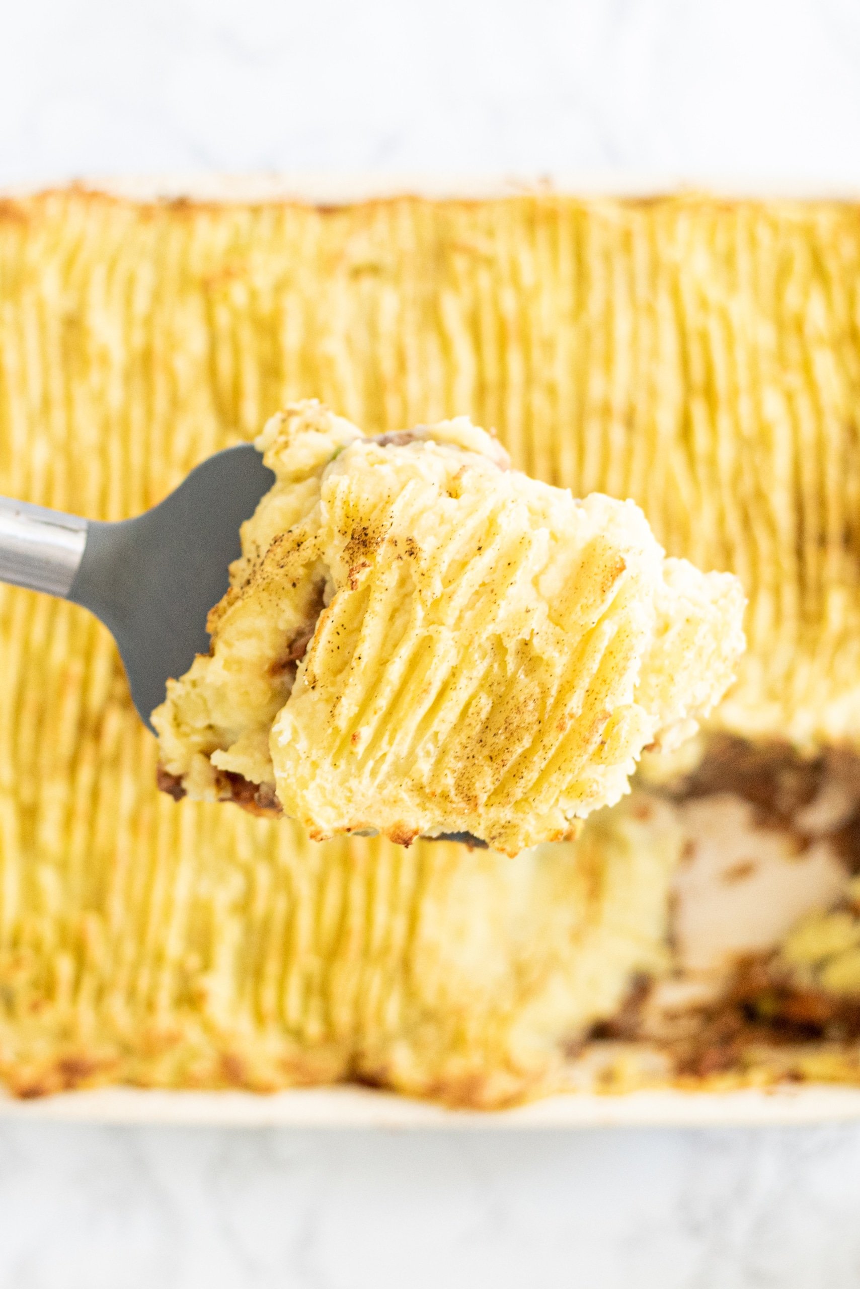 A casserole dish filled with shepherd's pie with a gray spatula holding a slice of pie.