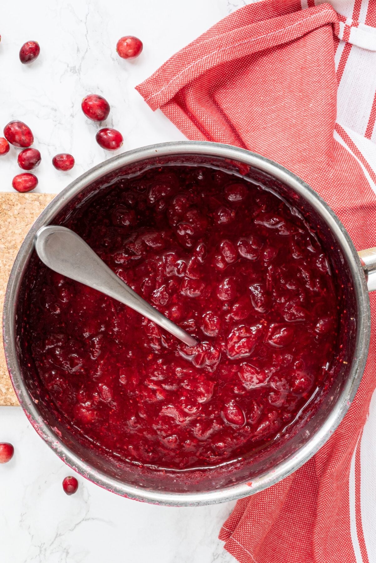 A silver pot of cranberry sauce with a silver spoon sticking out. Beside the pot is a white and red striped towel and some cranberries on the counter.