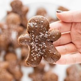 A white hand holding a gingerbread man cookie over a plate of remaining gingerbread cookies.