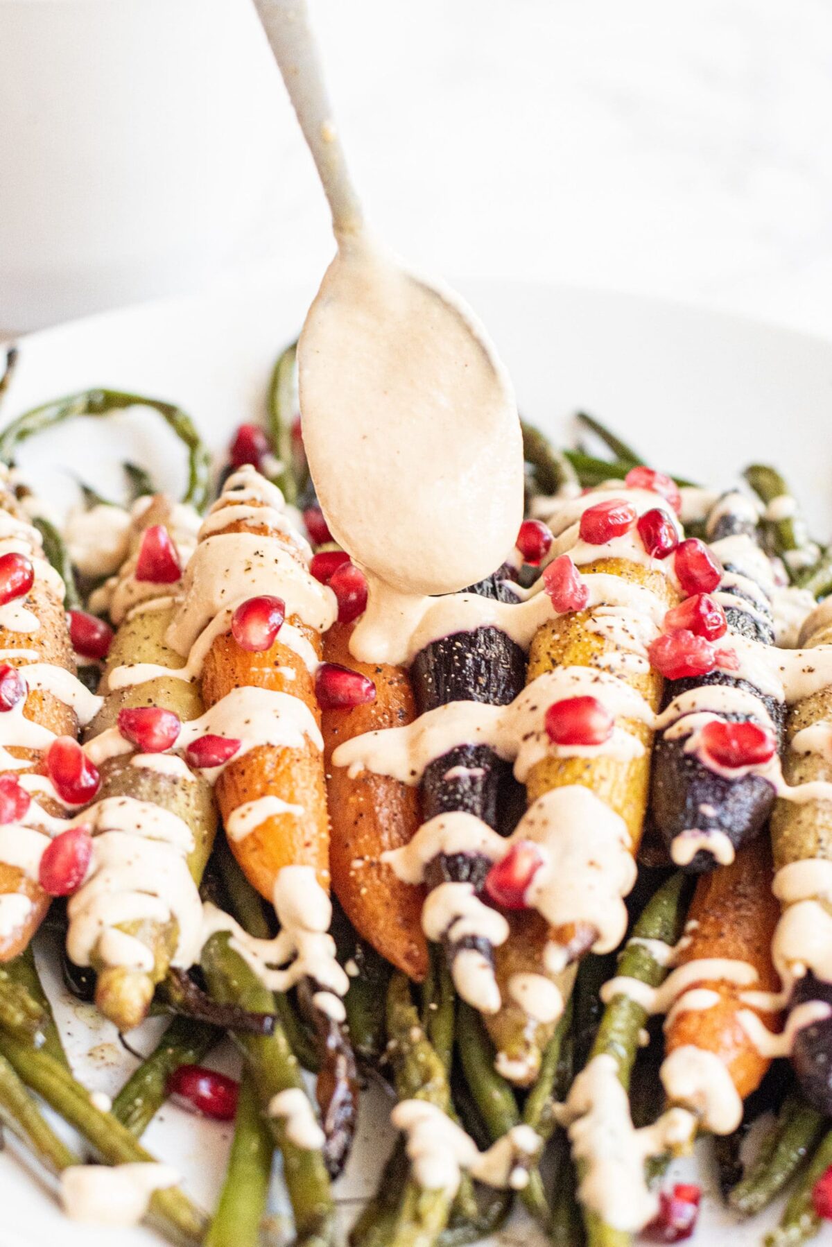A spoon with tahini on it and a plate filled with roasted carrots and green beans topped with tahini sauce and pomegranate seeds.
