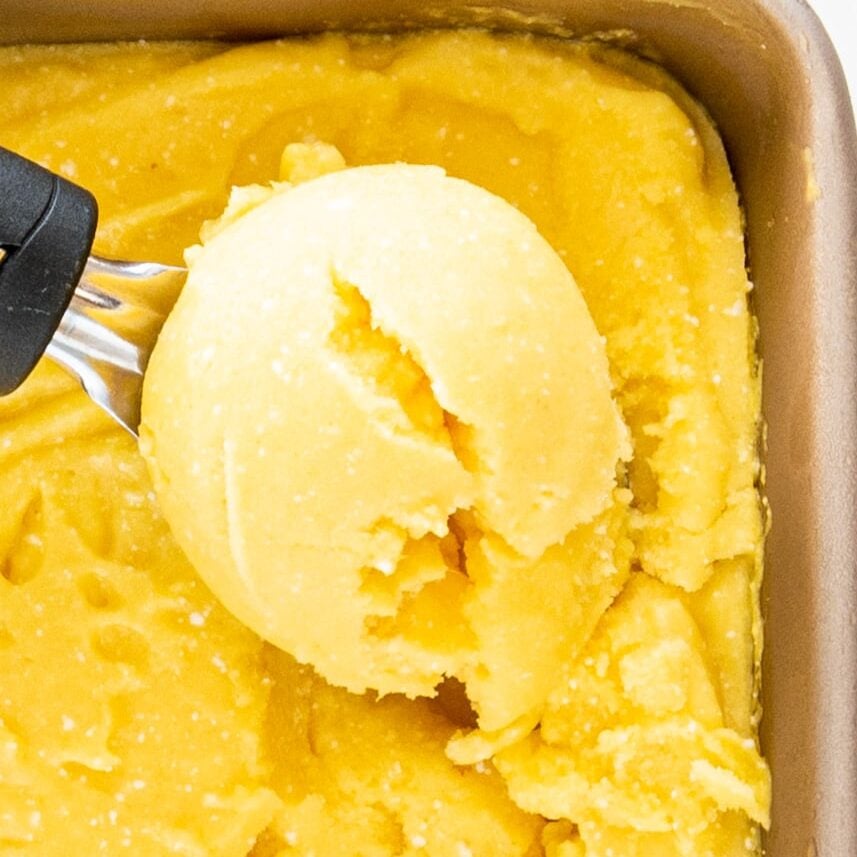 A metal pan filled with mango ice cream and a scooper filled with a scoop of ice cream.