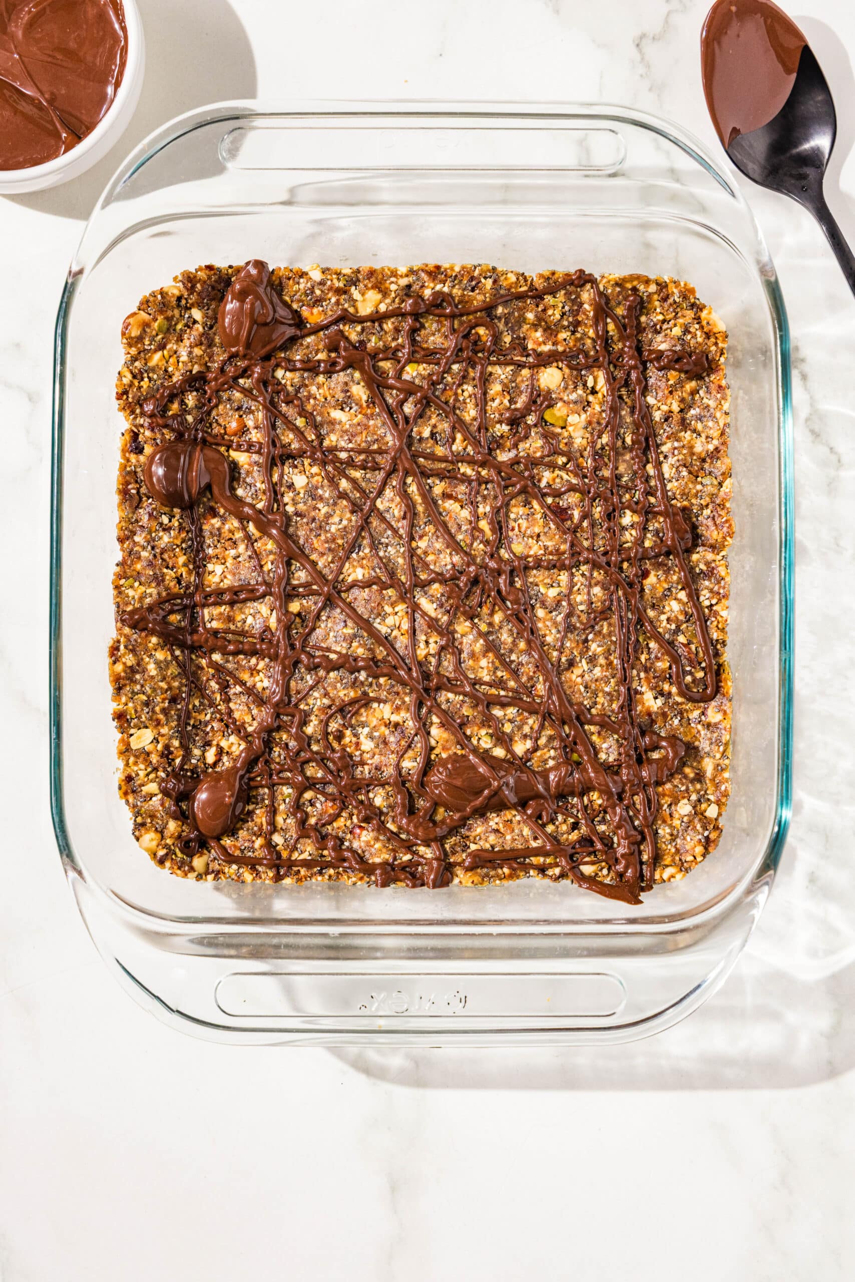 A glass 9x9 baking dish with a layer of vegan protein bars topped with a drizzle of chocolate. There is a small glass bowl filled with melted chocolate and a black spoon laying next to the baking dish.