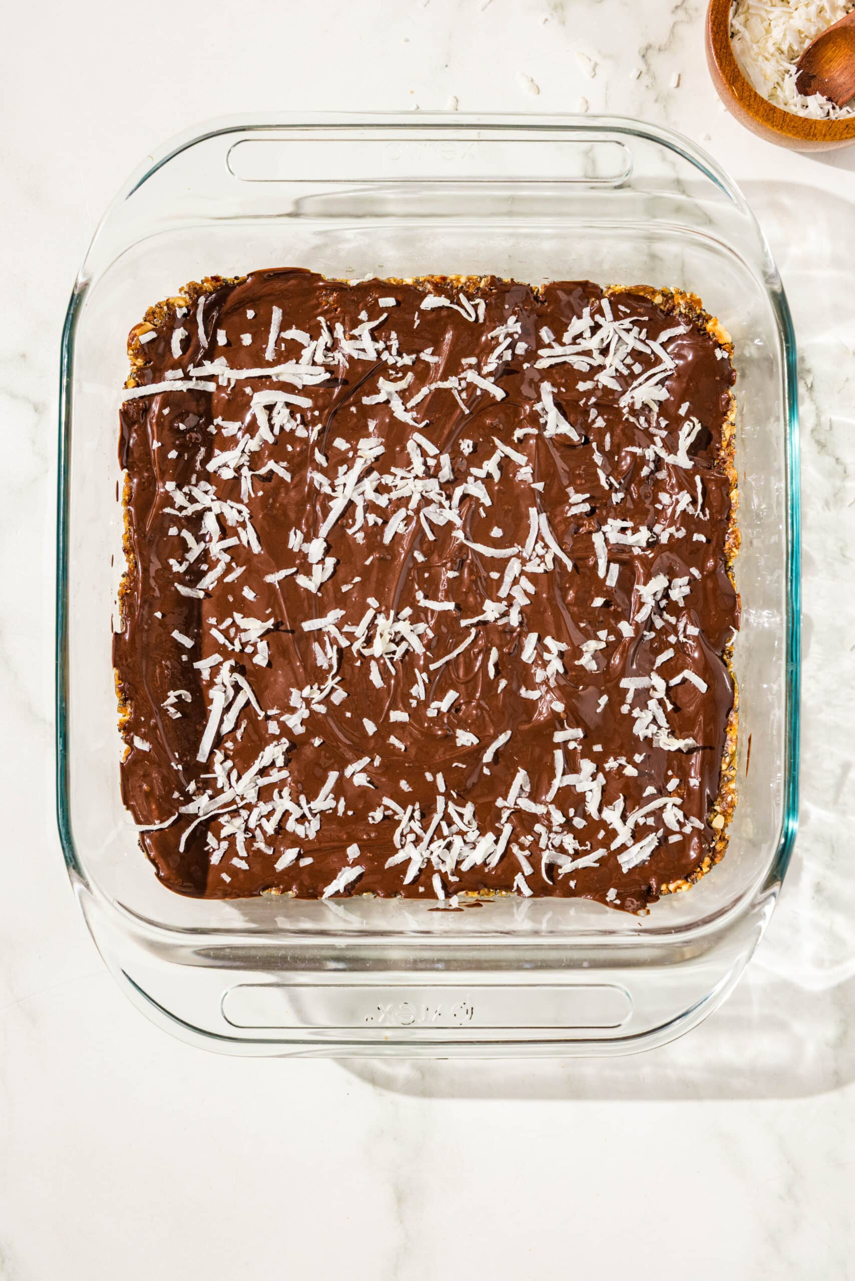 A glass 9x9 baking dish with a layer of vegan protein bars topped with a layer of chocolate and shredded coconut. There is a small wooden bowl filled with coconut next to the baking dish.