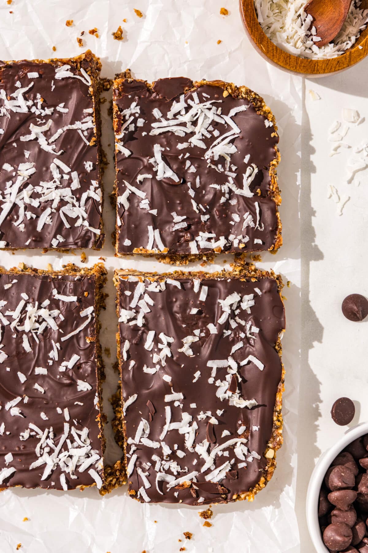 Four of the homemade vegan protein bars sliced on parchment paper with small bowls of chocolate chips and shredded coconut surrounding.