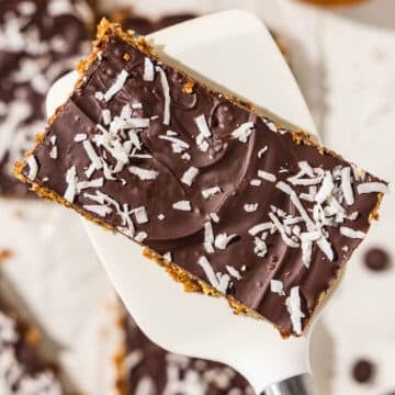 A homemade vegan protein bar with melted chocolate and shredded coconut on top sitting on top of a spatula. In the background there are more protein bars and a small wooden bowl filled with coconut.