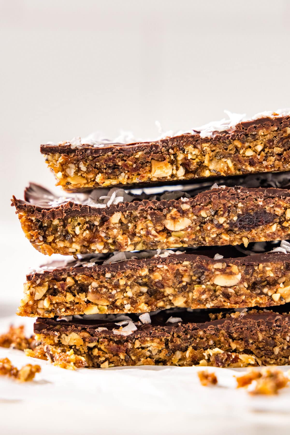 A stack of four homemade vegan protein bars, each coated in dark chocolate and shredded coconut on the top. There is crushed nuts surrounding the bars.