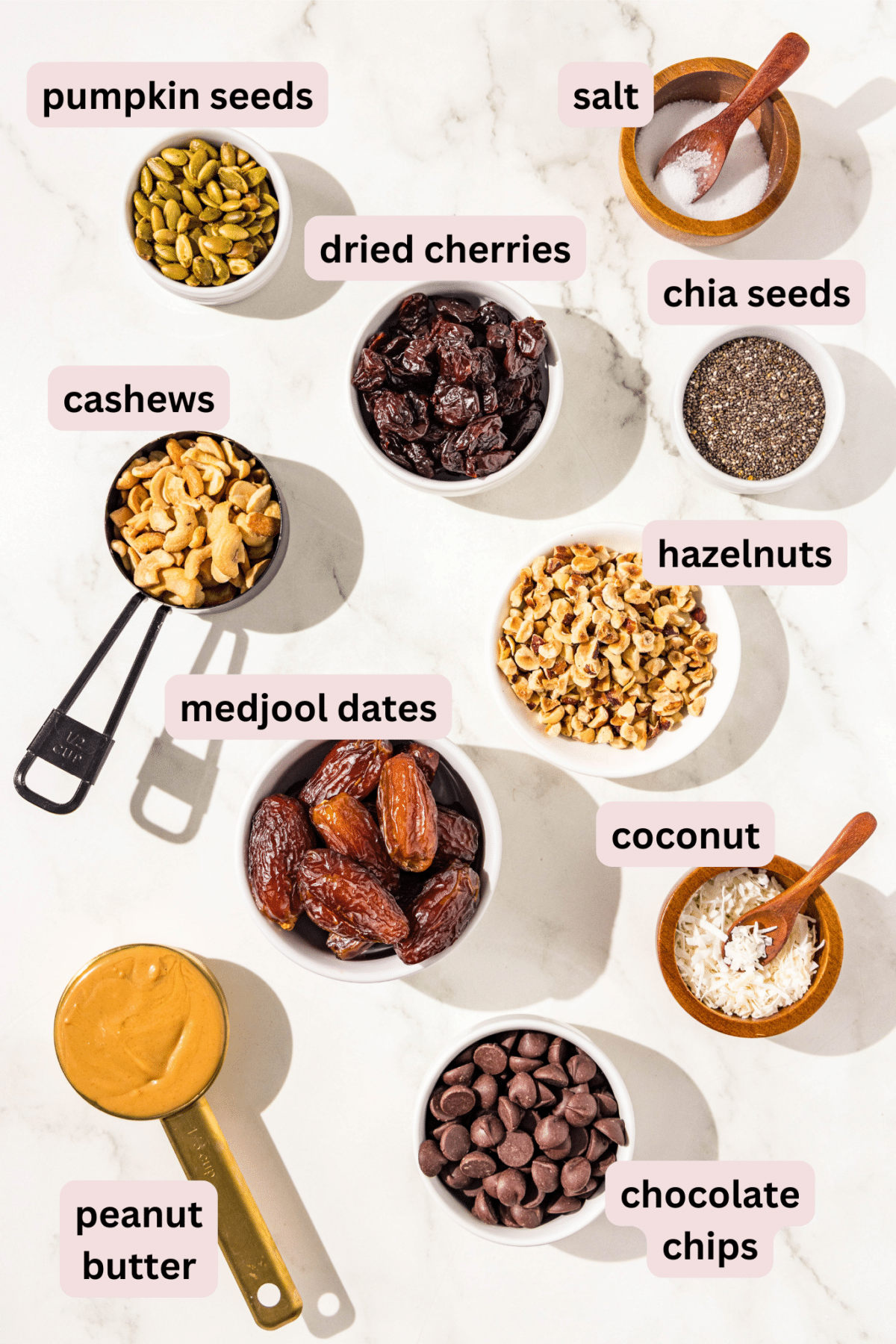 A recipe ingredients image showing ingredients in small bowls including salt, pumpkin seeds, dried cherries, chia seeds, cashews, hazelnuts, medjool dates, coconut, peanut butter, and chocolate chips.
