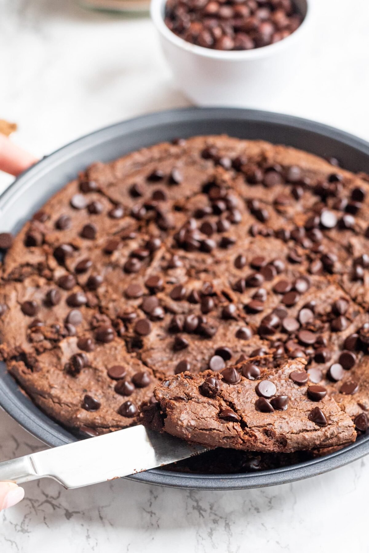 A round pan filled with vegan black bean brownies and a silver knife cutting into them. In the background is a white hand holding the pan and a small white bowl of chocolate chips.