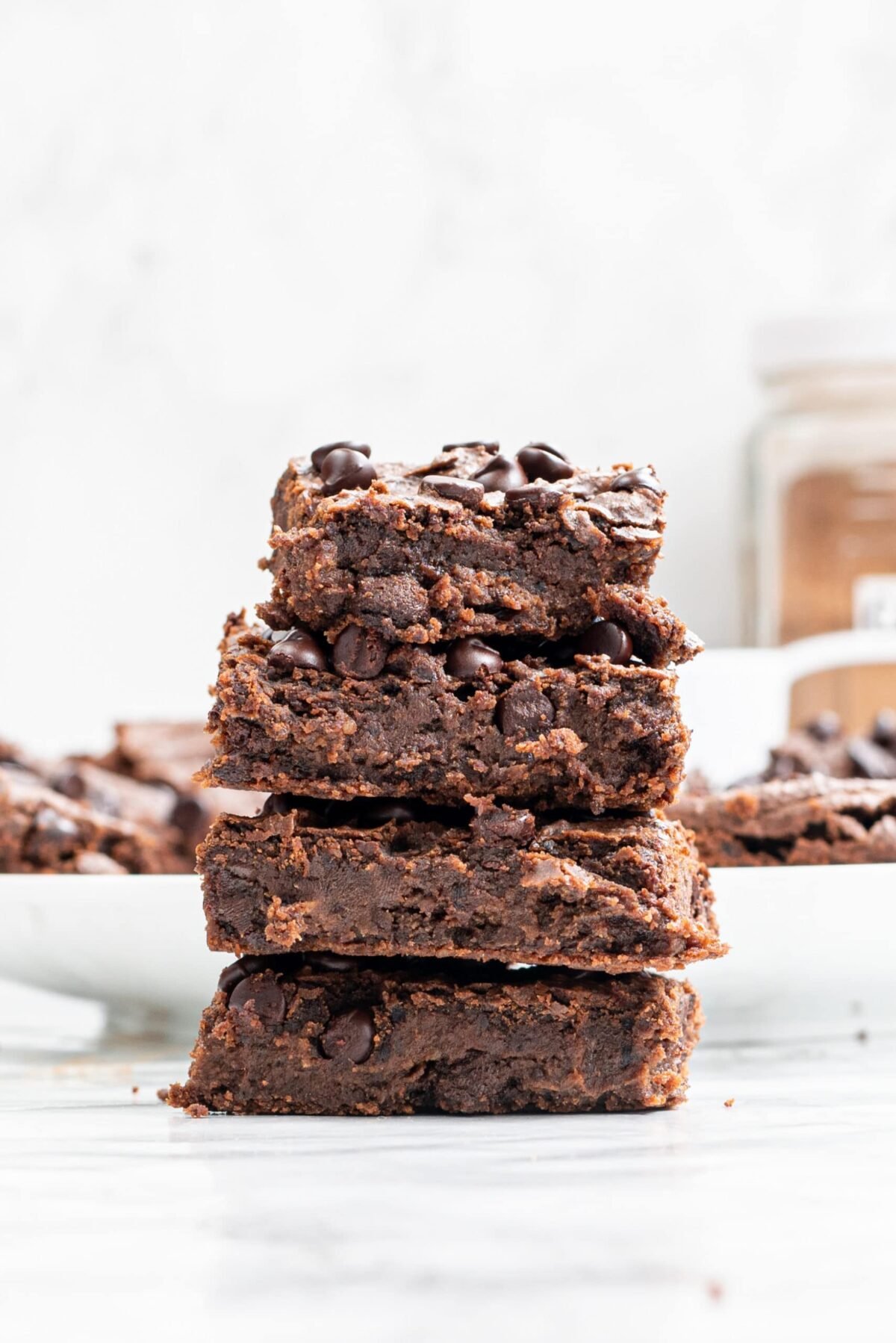 Four Vegan Black Bean Brownies stacked on top of each other with more brownies in the background on a white plate and a glass jar of cacao powder.