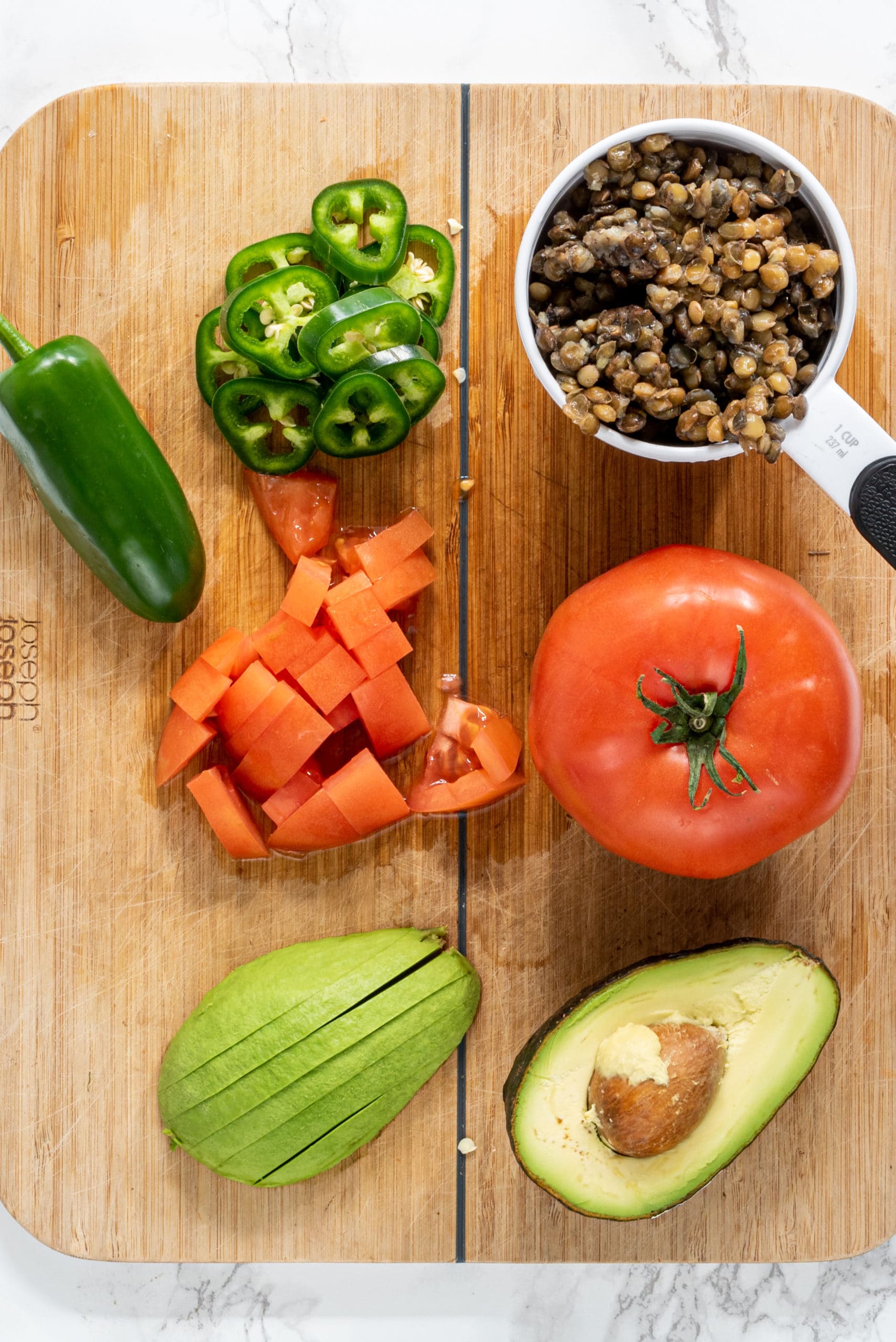 A wood cutting board with a halved sliced avocado, a whole tomato and a sliced tomato, a cup of lentils, and sliced jalapenos.